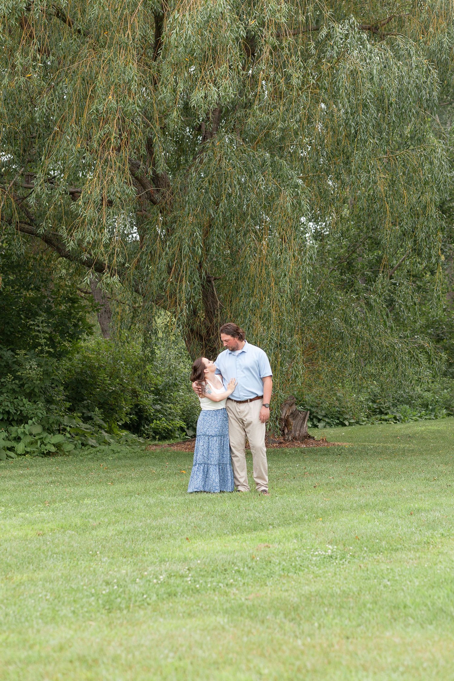 Romantic couple embraces under a graceful weeping willow tree in a serene garden setting.