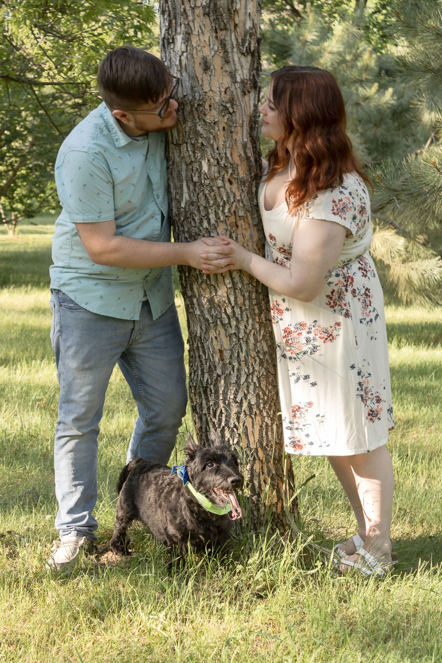 Couple poses playfully around a tree trunk with their small black dog in a sunny garden setting.
