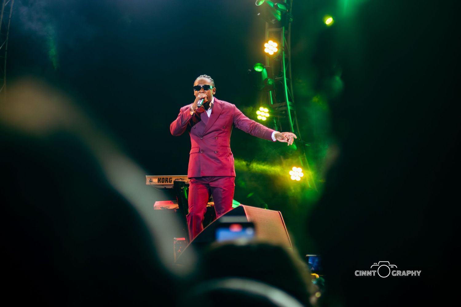 A performer in a red outfit jumps energetically on stage during a concert with green lighting effects.