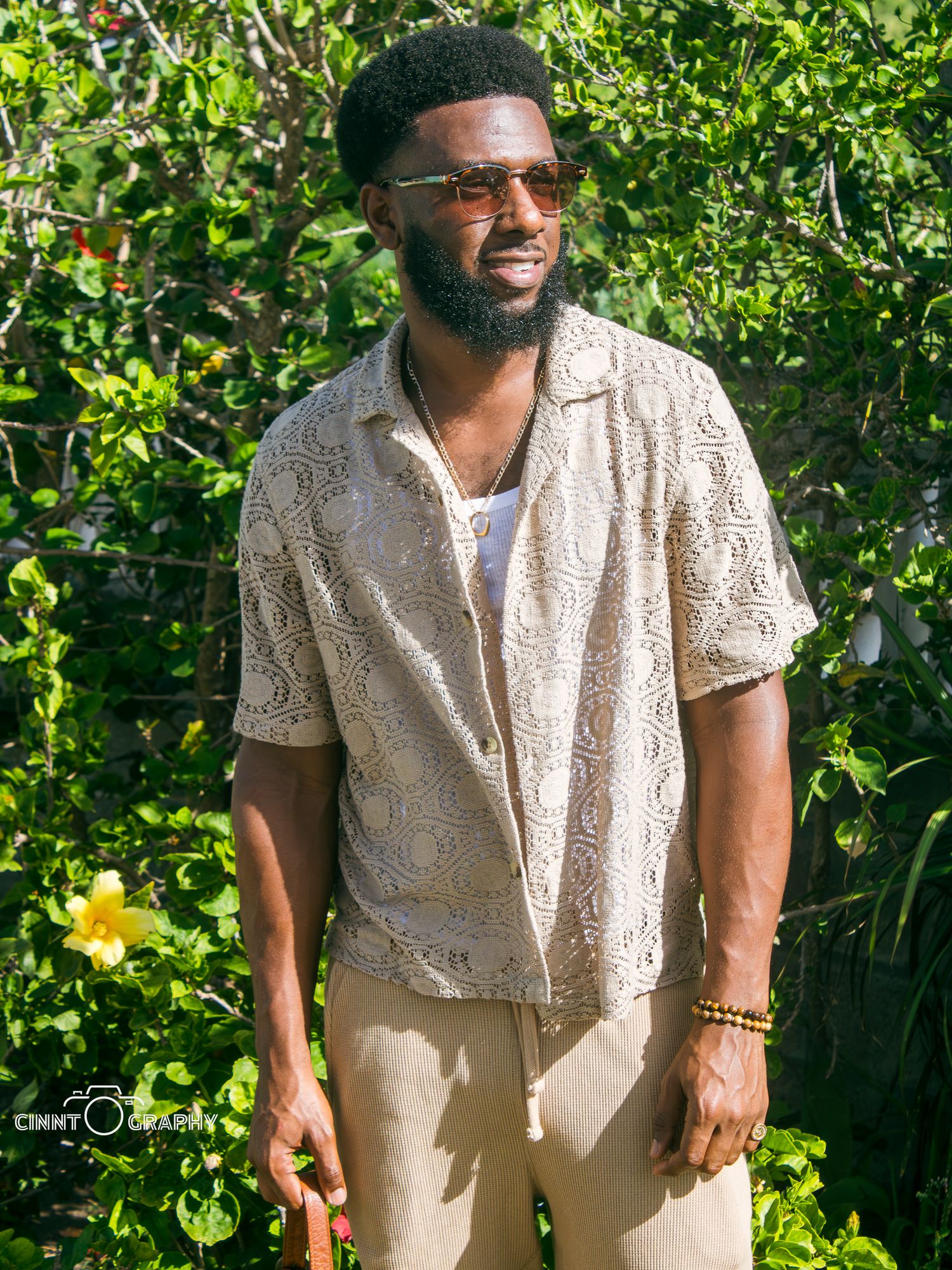 Person in light-colored summer attire poses playfully in a lush garden setting with vibrant green foliage in the background.