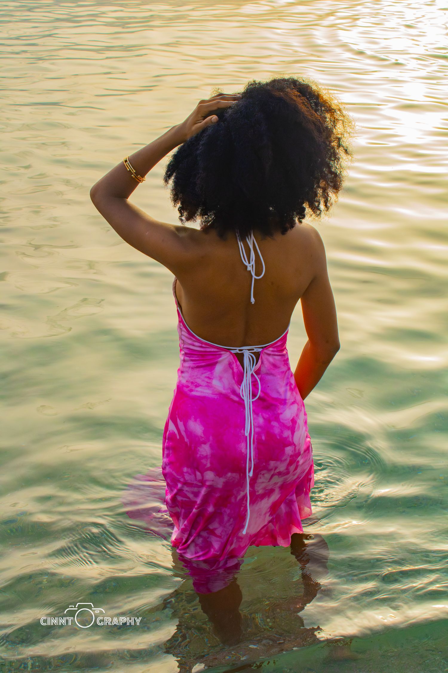 View from behind of person in pink tie-dye dress standing in reflective water during golden hour.