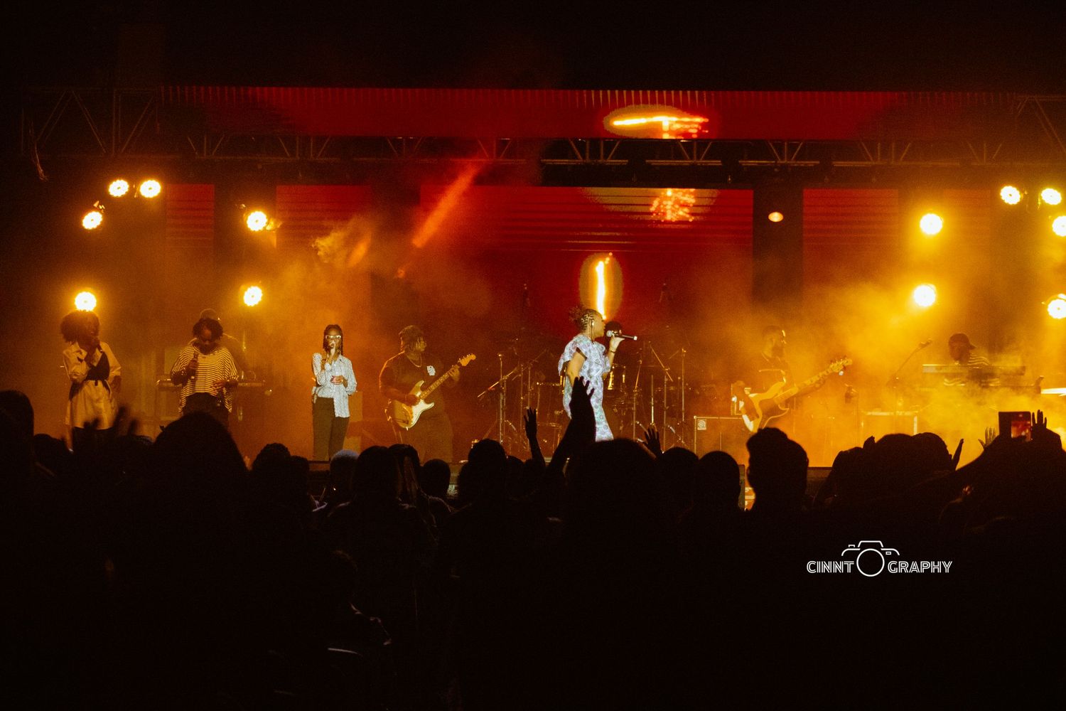 A silhouetted performer and band on stage bathed in dramatic orange and red concert lighting with crowd in foreground.