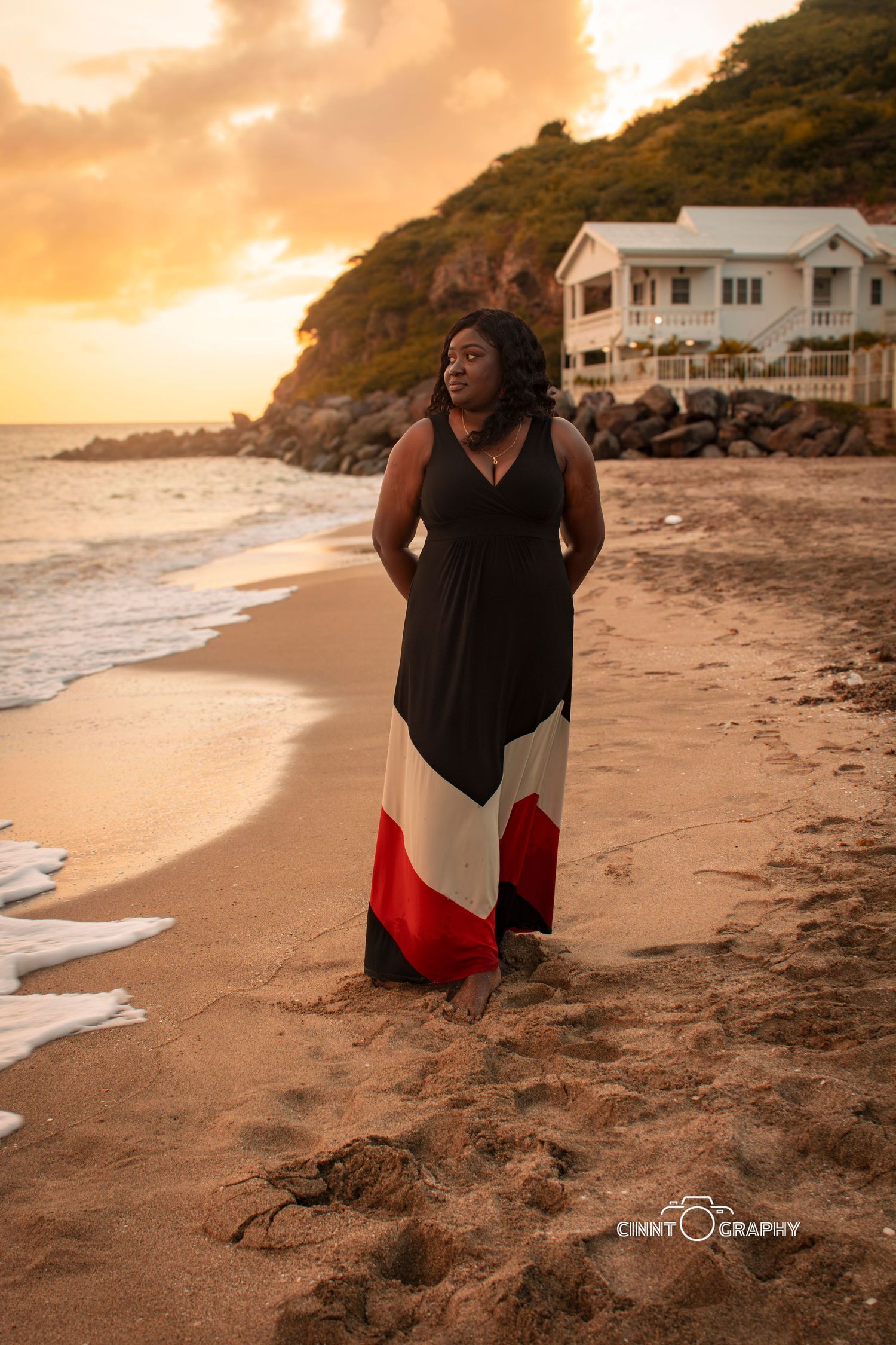 Woman in black top looks thoughtfully towards sunset on sandy beach with waves and coastal rocks in the background.