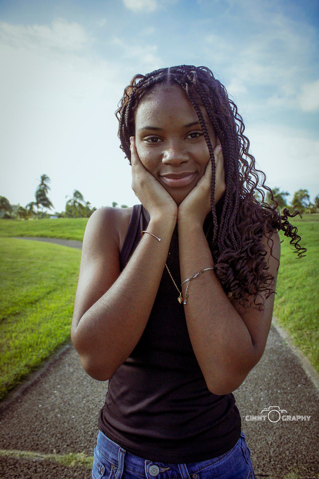 A smiling person with curly hair poses outdoors on a path with green grass and blue sky in the background.