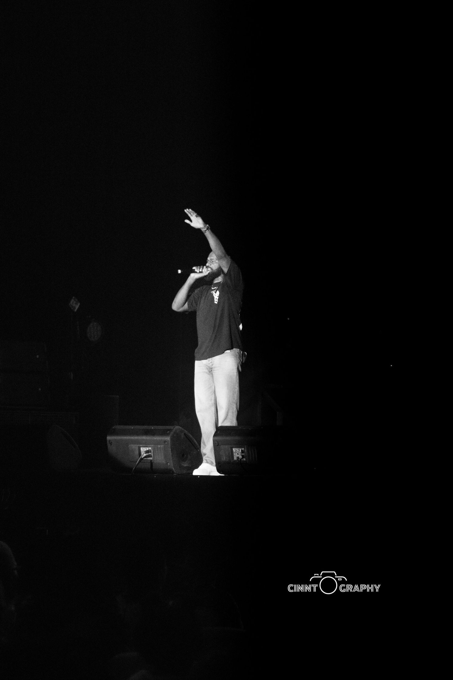Performer in white pants and dark shirt stands on stage with raised arm in dramatic black and white spotlight.
