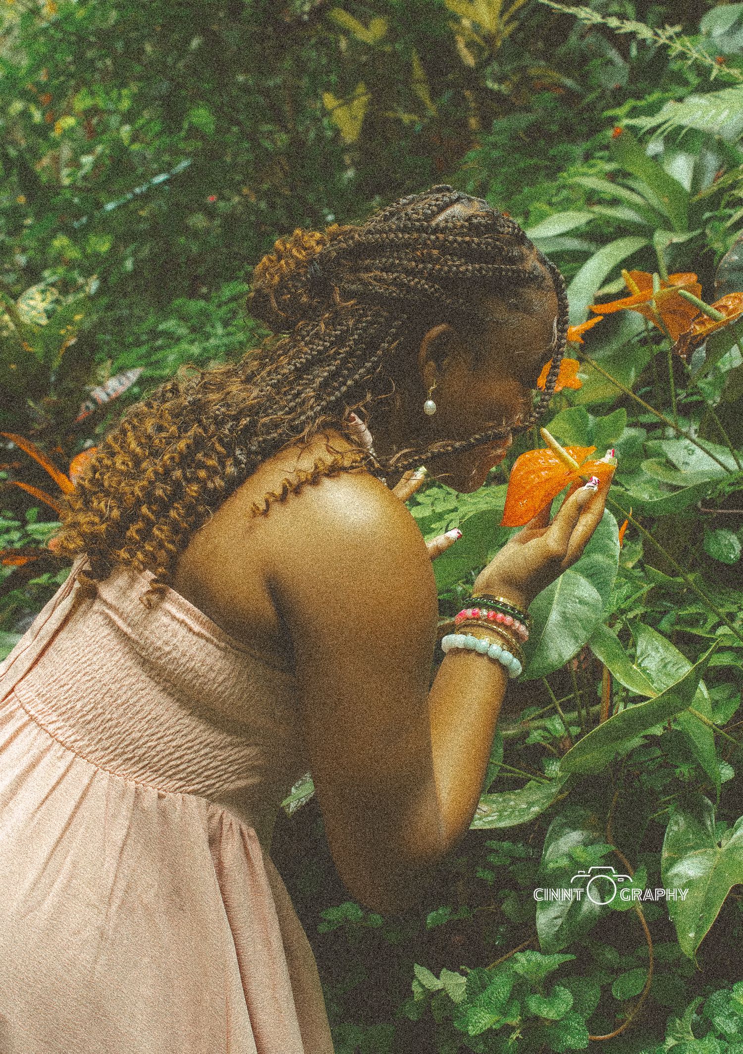 Someone in a beige dress with braided hair leans forward to smell bright orange flowers in a lush garden setting.