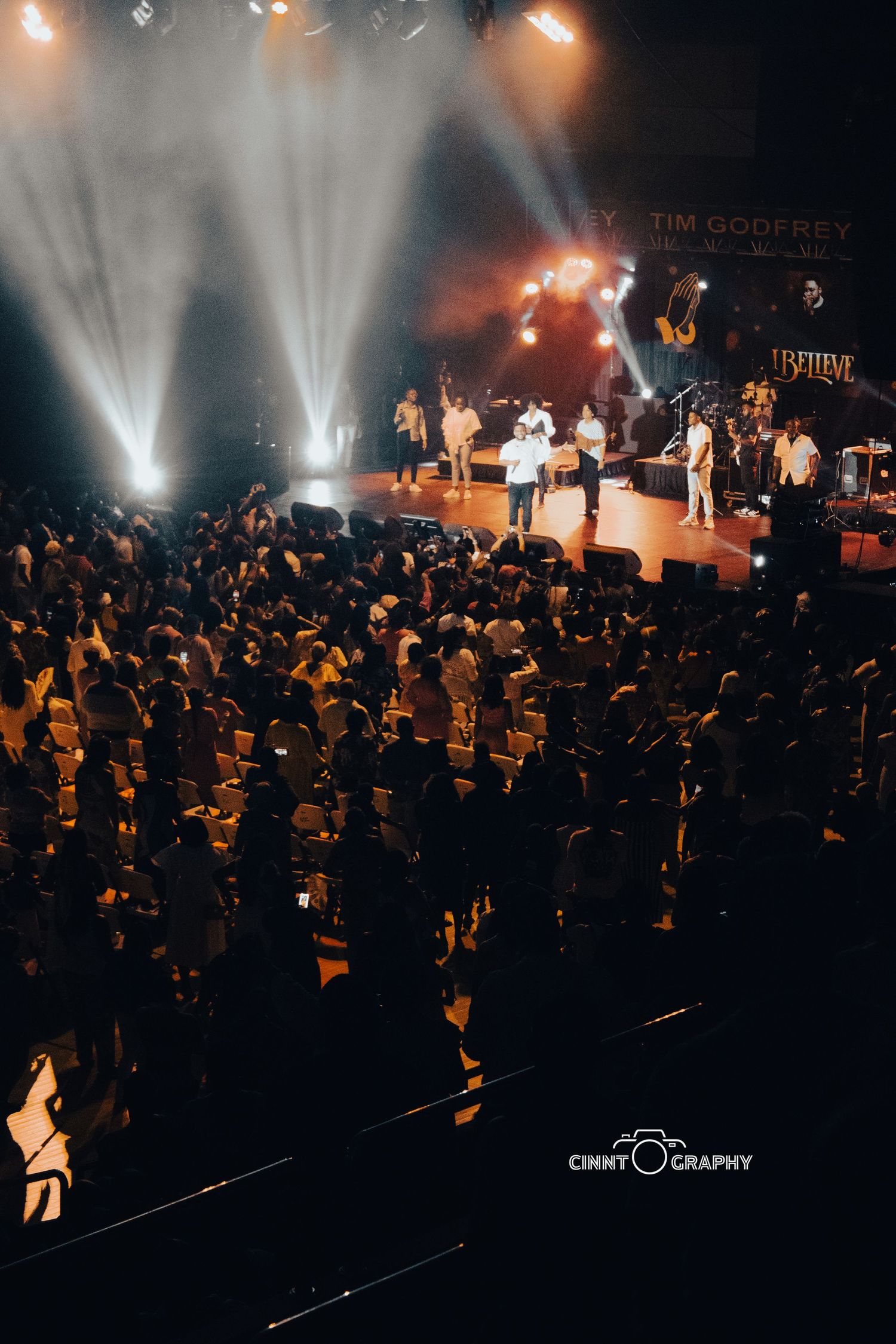 Concert performers on stage illuminated by bright spotlights with a large audience crowd silhouetted below at a music venue.
