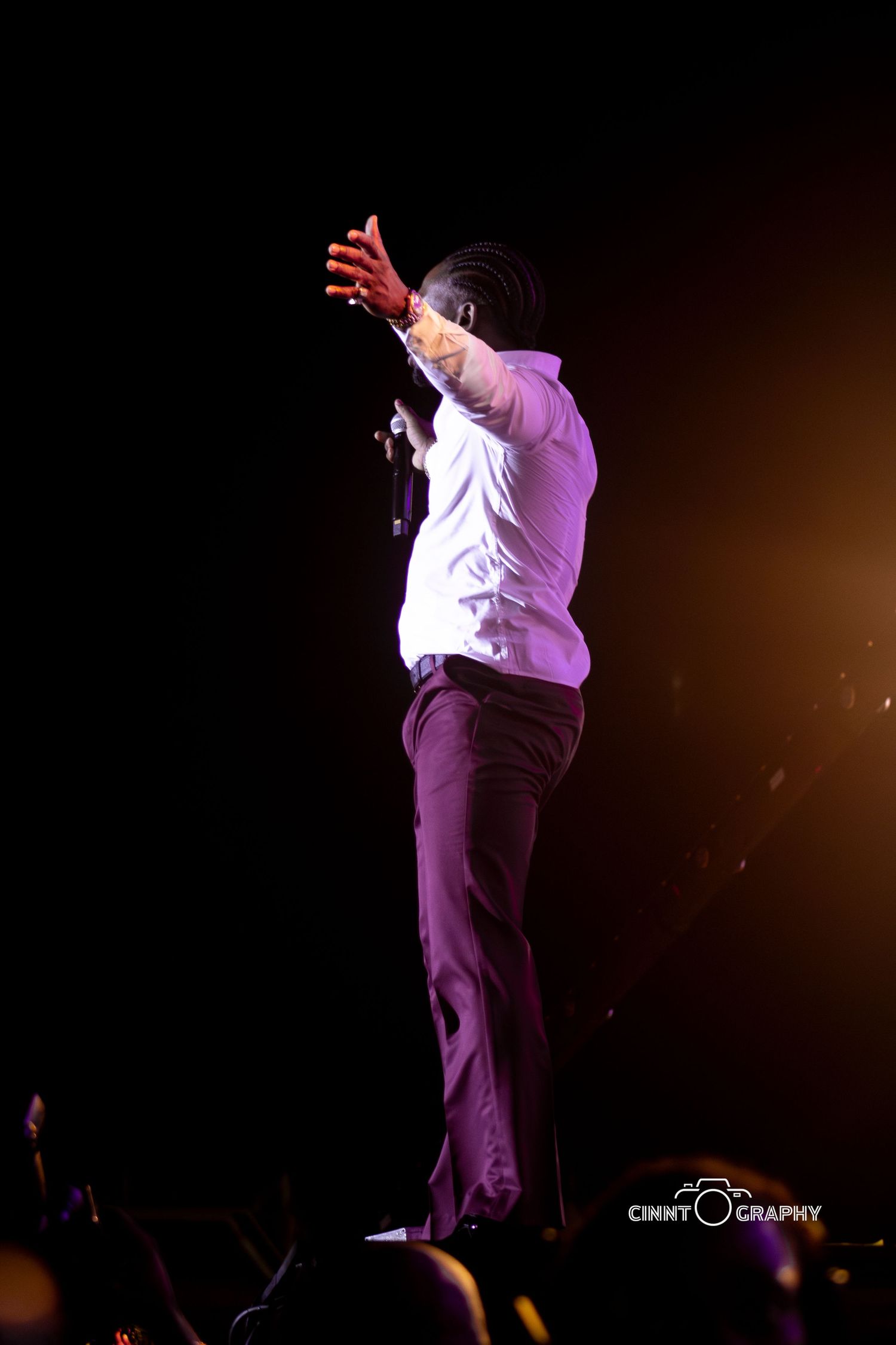 Concert performer in white shirt and dark pants gestures dramatically under purple stage lighting.