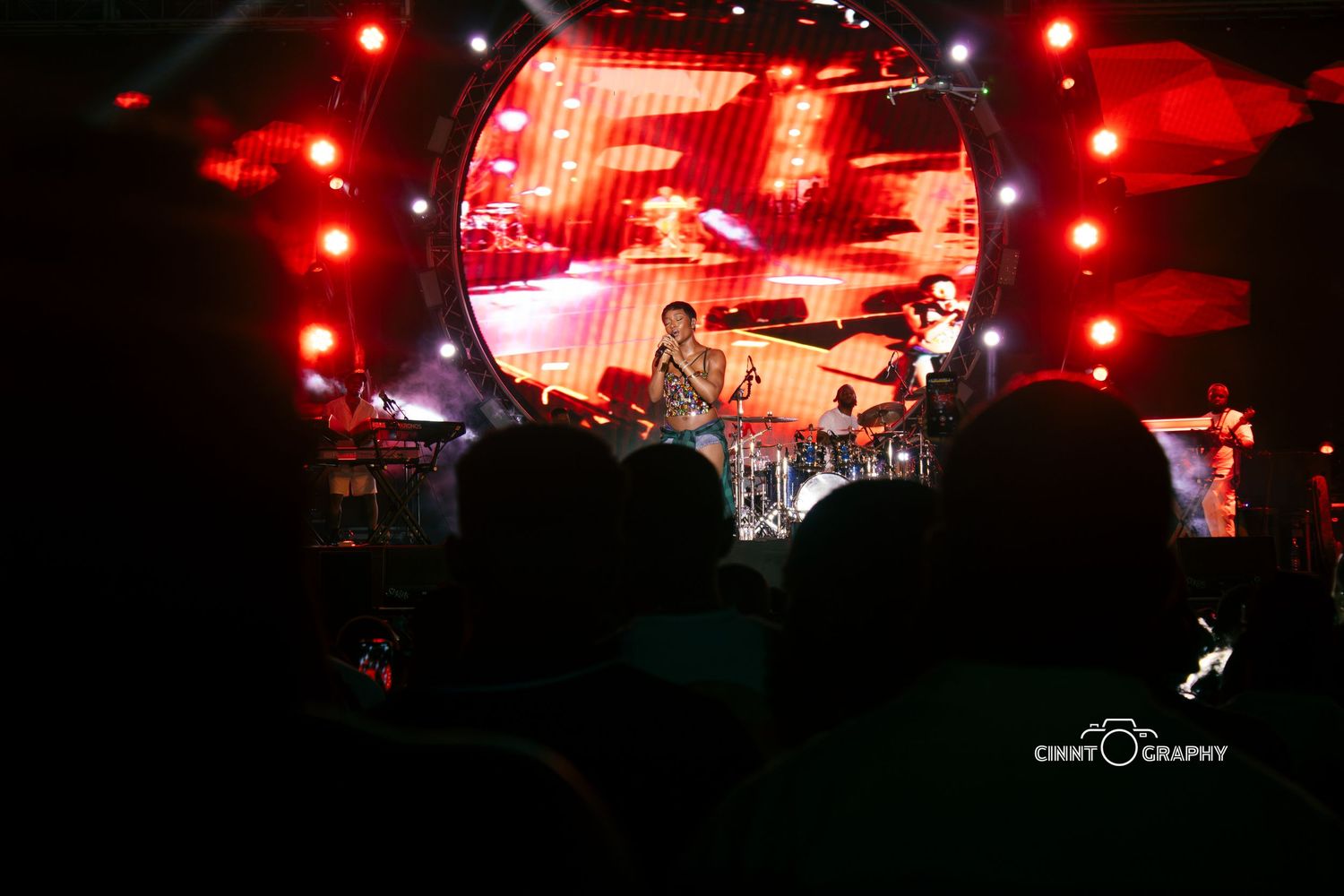 Concert stage lit with dramatic red lighting and video screens displays vibrant performance atmosphere.