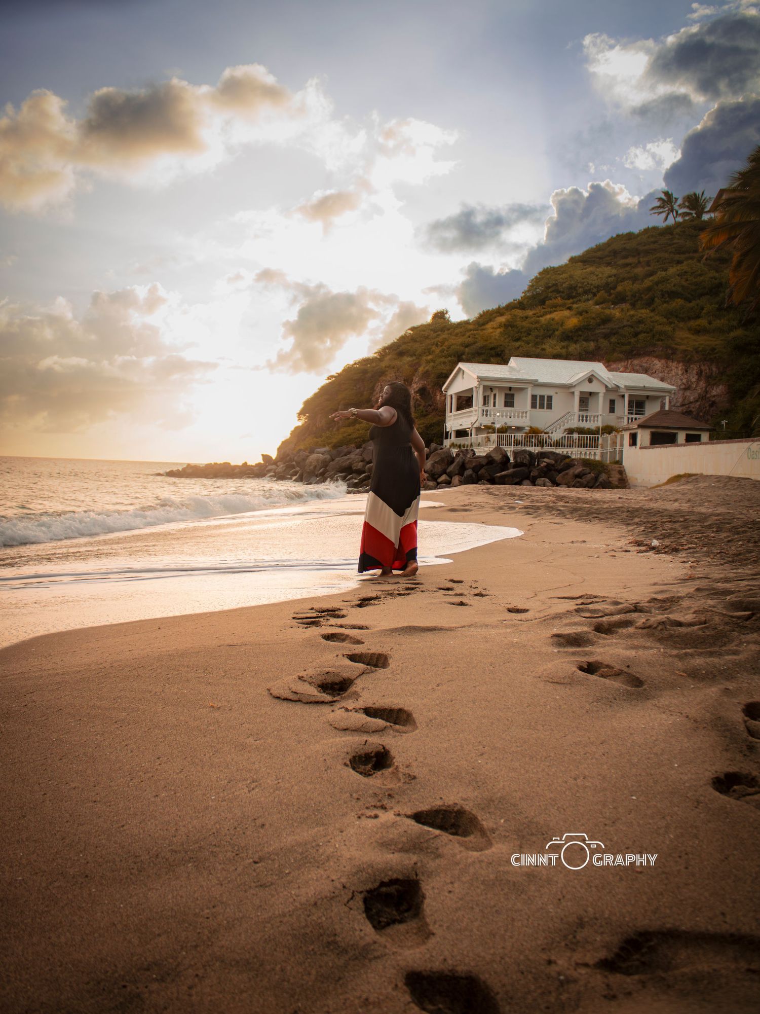 Figure in flowy skirt stands on beach at sunset with footprints in sand leading to beachfront house on cliff.