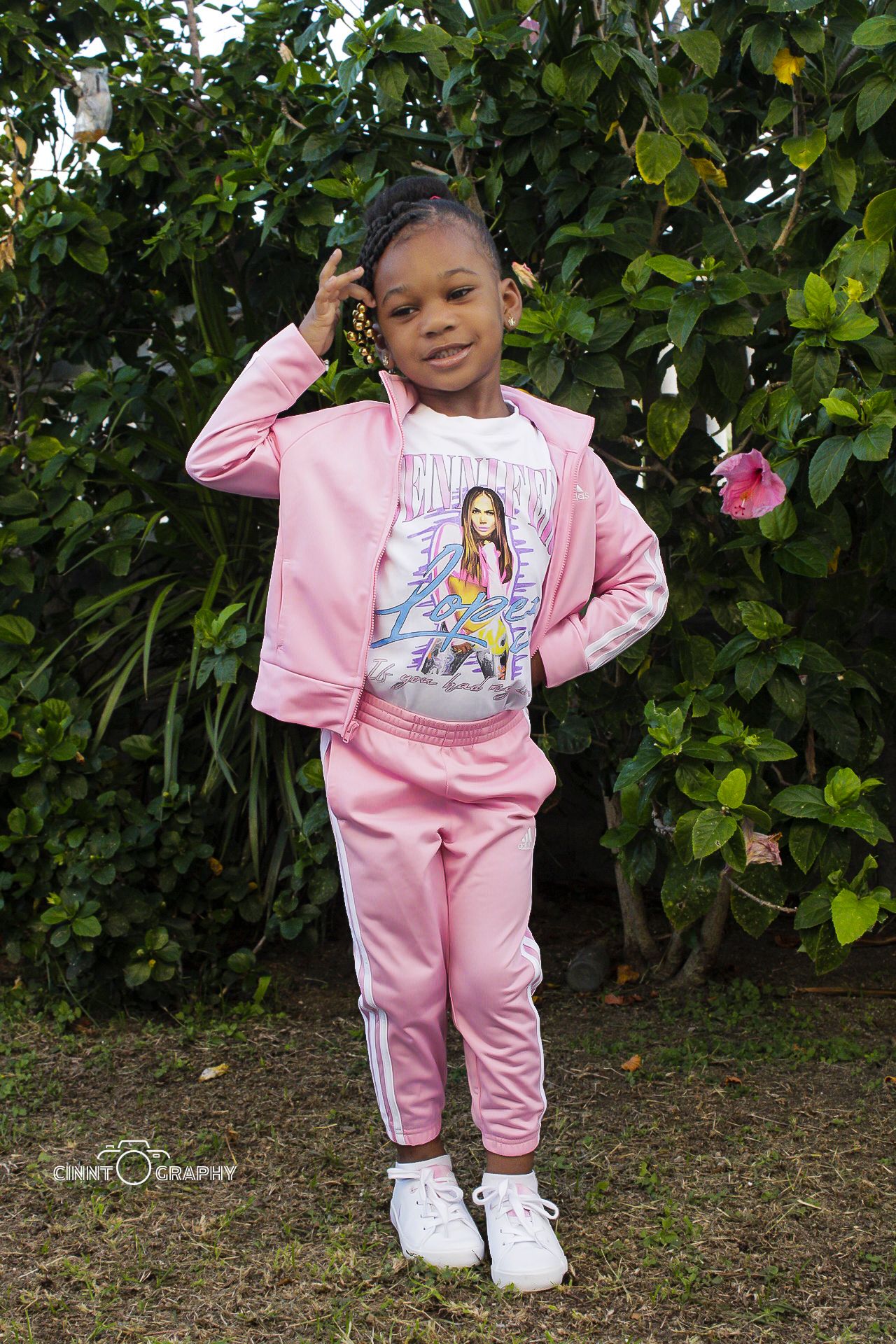 A young child poses in a matching pink tracksuit and white sneakers against a leafy green garden background.