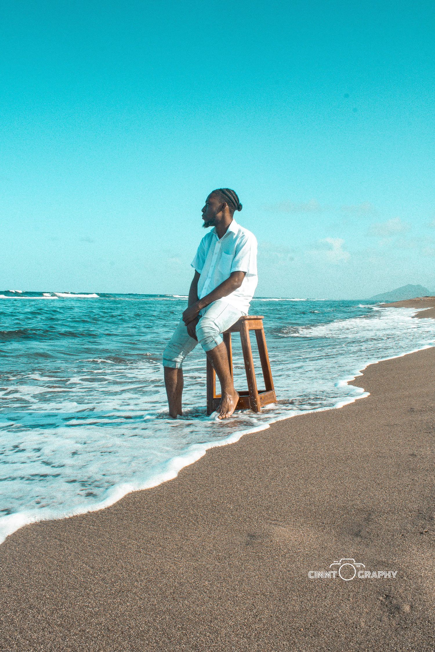 Person sitting on wooden stool at beach edge with turquoise ocean waves and clear blue sky in background.