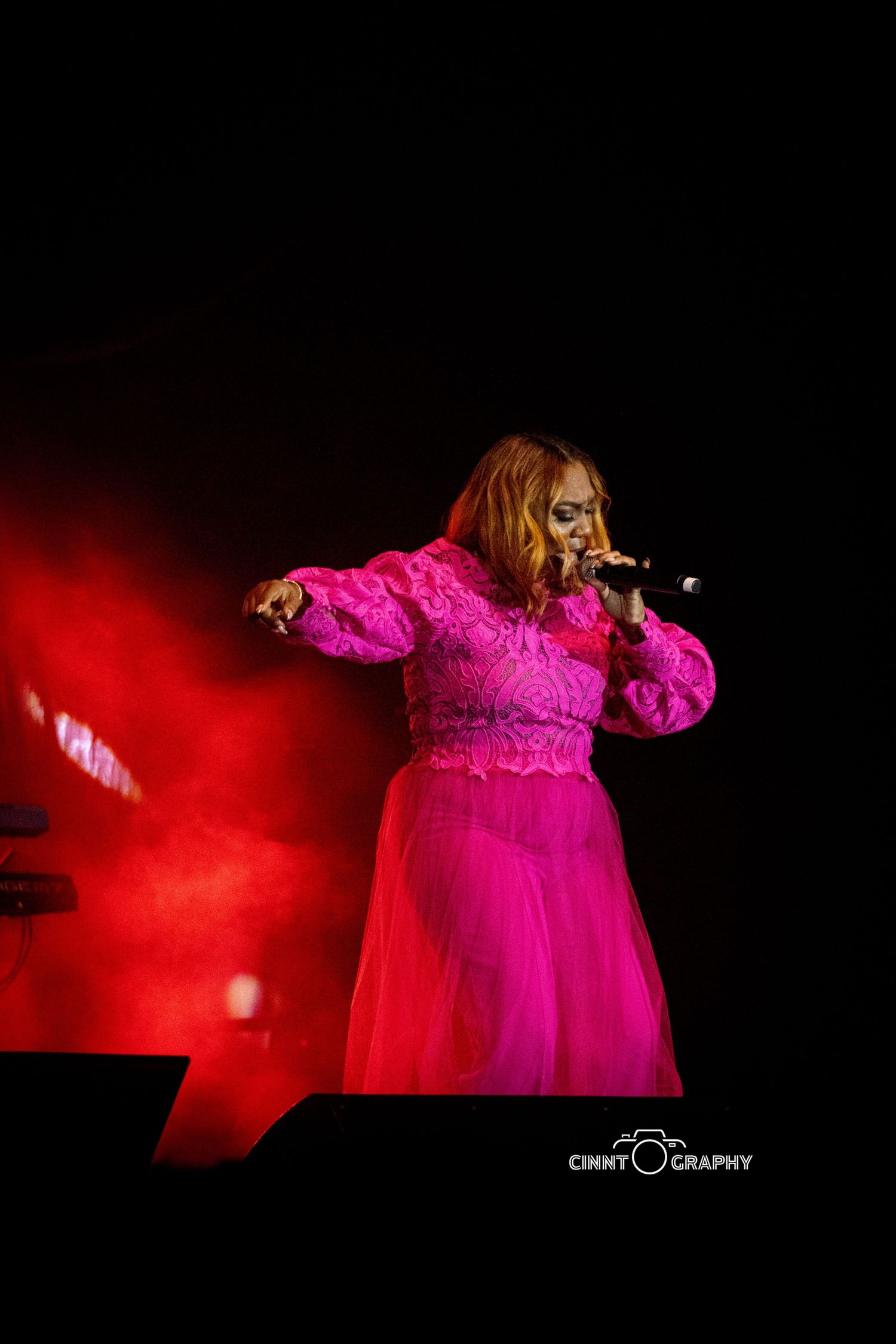 Performer in a bright pink lace gown singing passionately on stage amid dramatic red lighting effects.