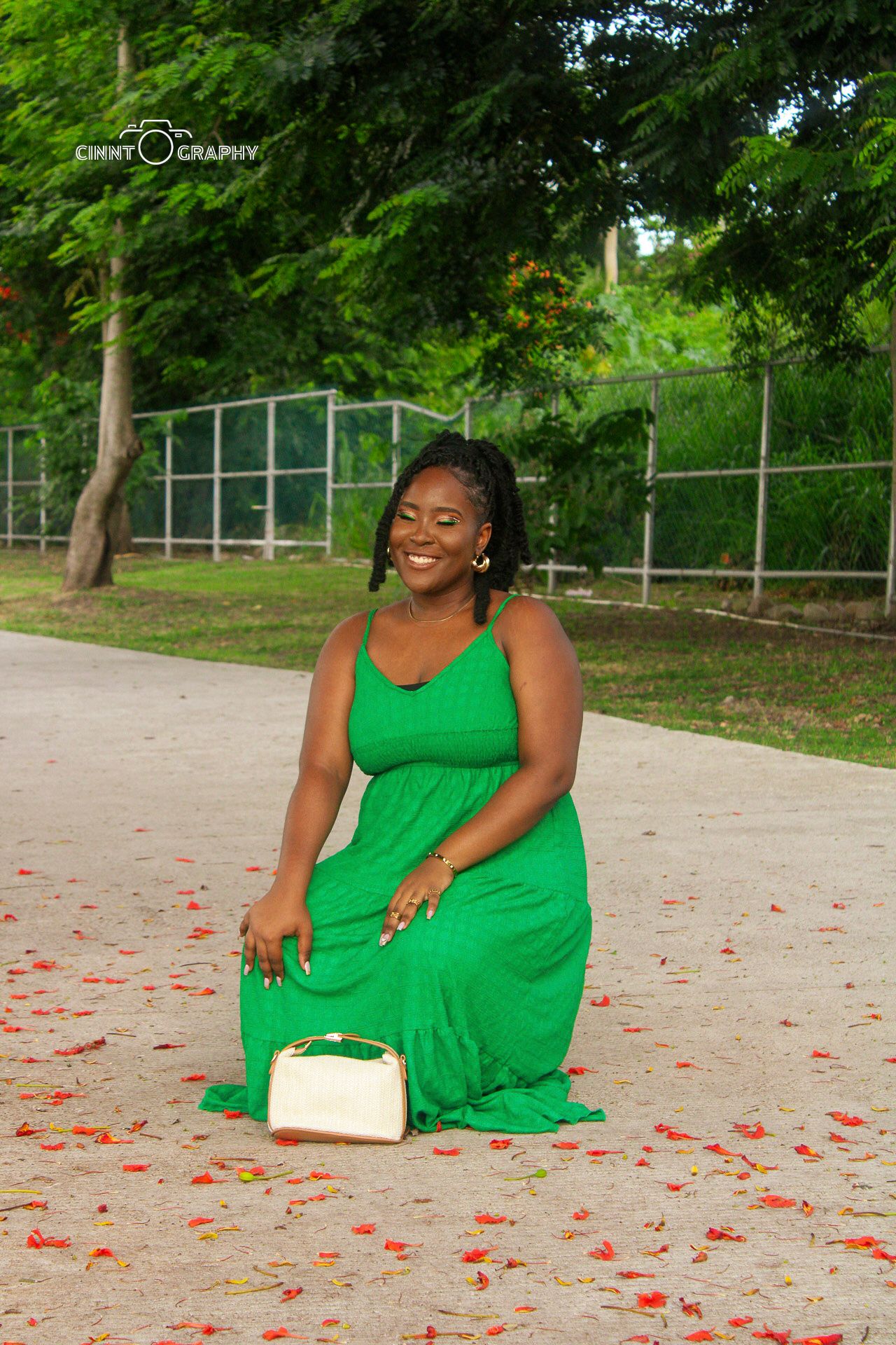 A woman in a vibrant green maxi dress sits gracefully on the ground with scattered red petals and a light-colored purse.