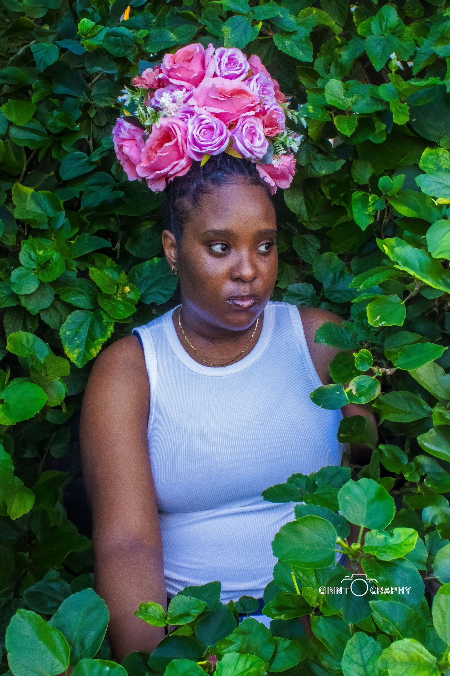 Pink floral crown adorns figure surrounded by lush green foliage in artistic garden portrait.