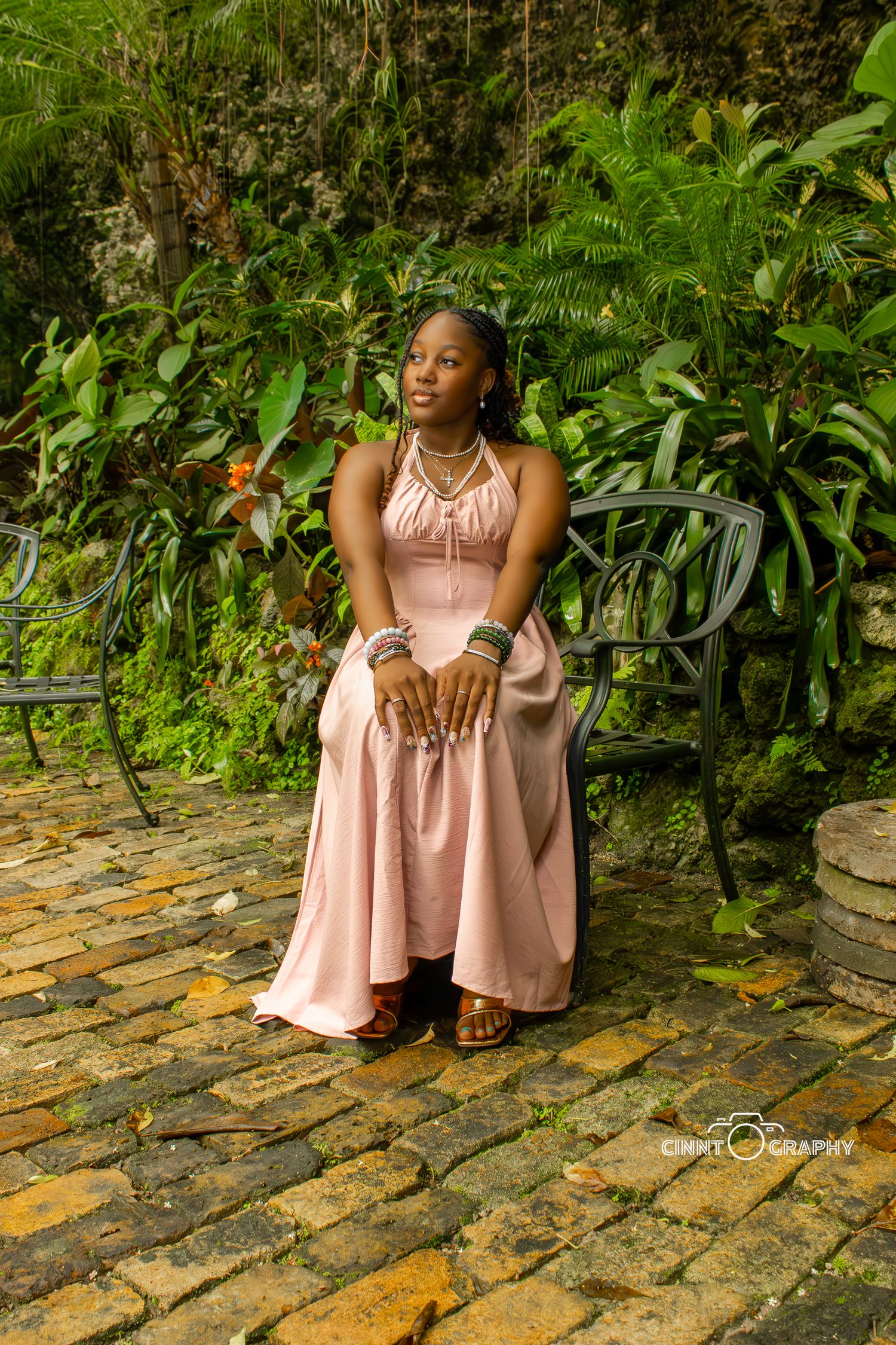 A person in a pink dress sits thoughtfully on stone steps surrounded by lush tropical greenery.