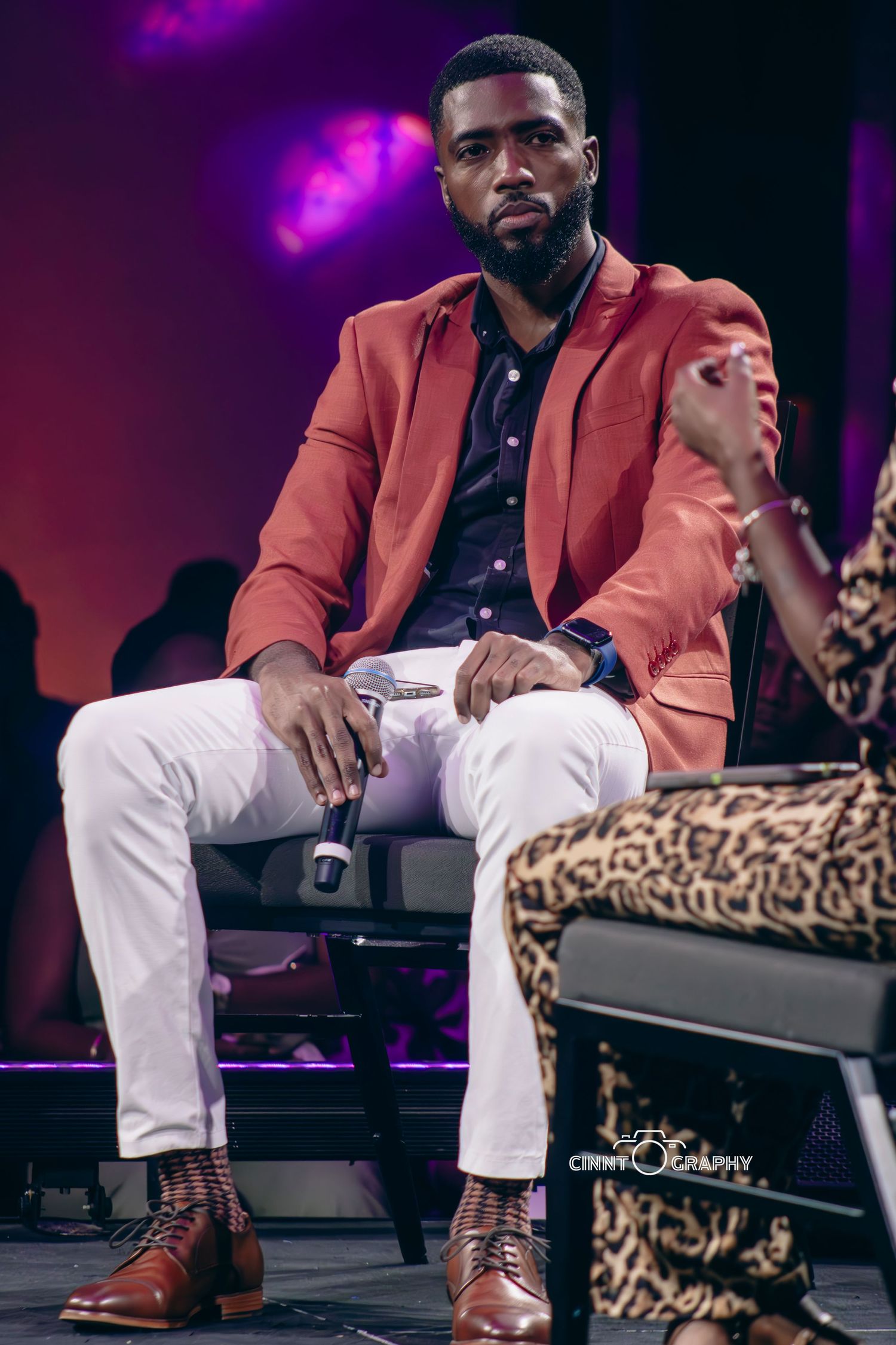 Stylish portrait in salmon blazer and white pants against purple stage lighting.