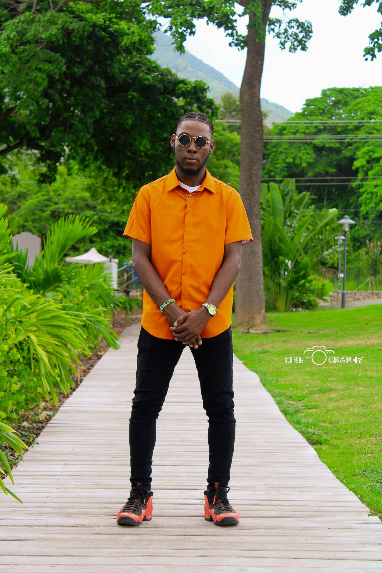 A person in an orange button-down shirt and black pants poses on a wooden walkway surrounded by lush tropical landscaping.