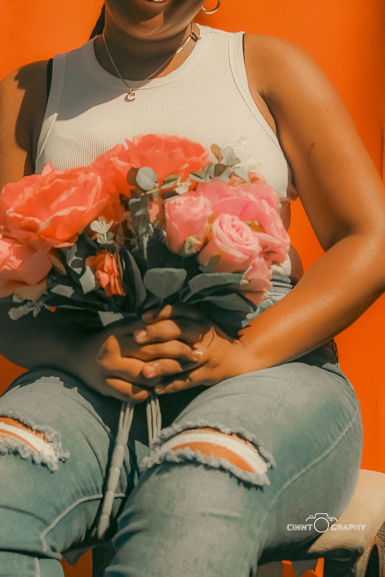 Someone wearing a white tank top and ripped jeans holds a vibrant bouquet of pink and coral roses against an orange background.