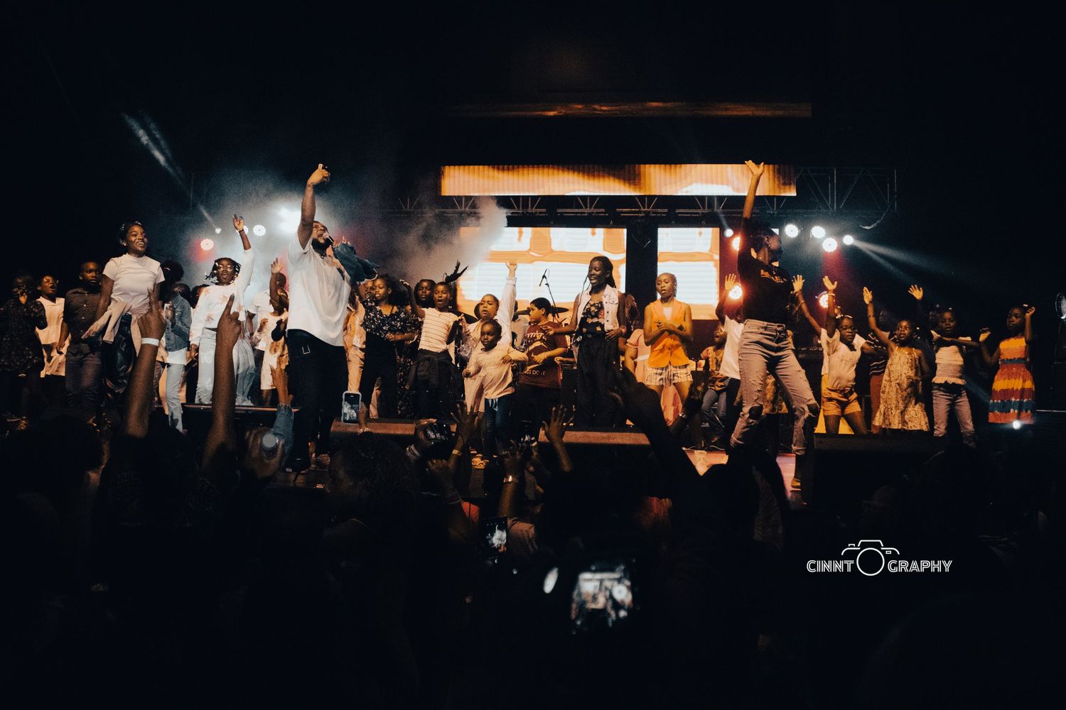 A crowd of people with raised hands enjoying a lively concert performance on stage illuminated by warm lighting at night.