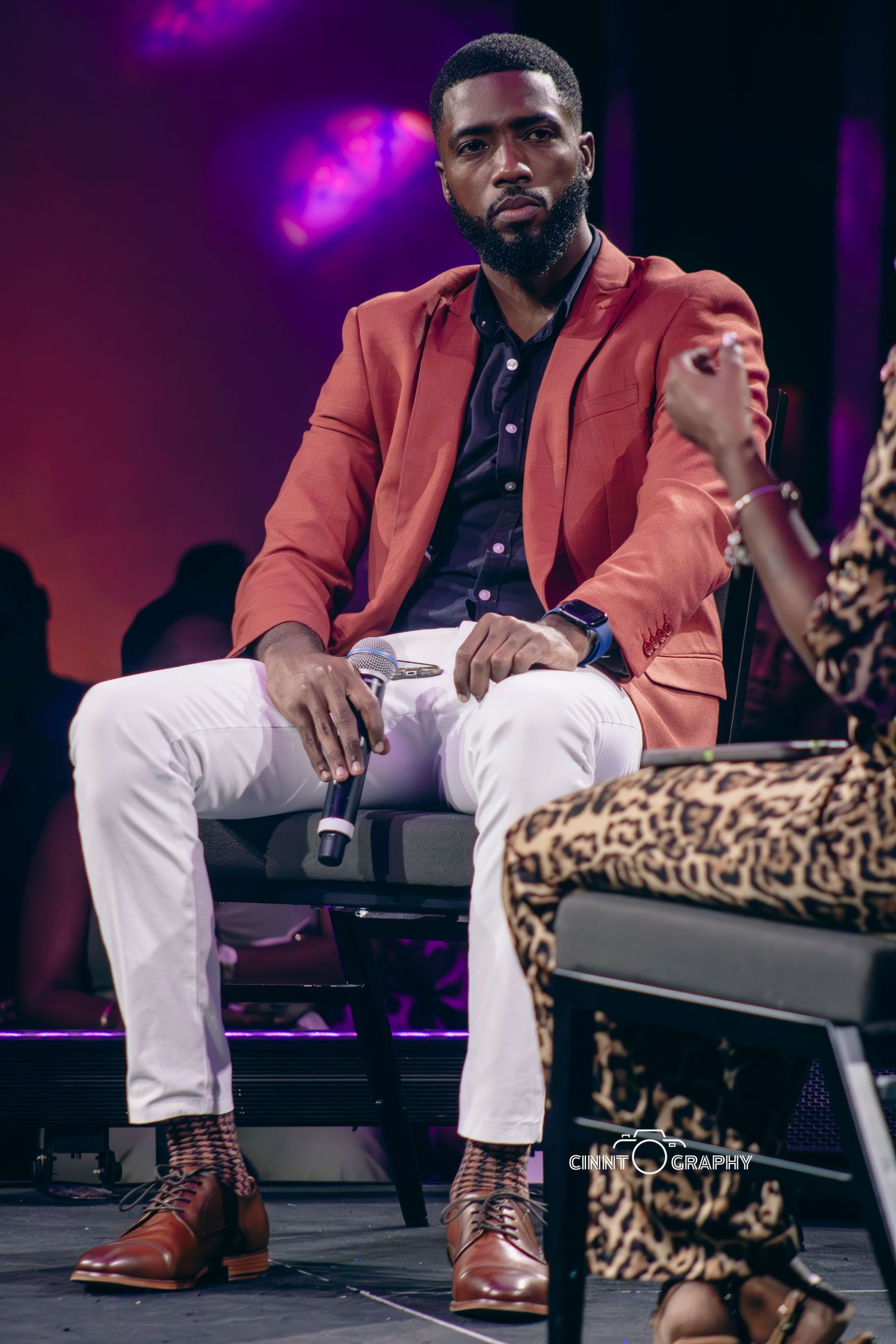 Stylishly dressed person in salmon-colored blazer and white pants sits on stage with purple lighting effect.