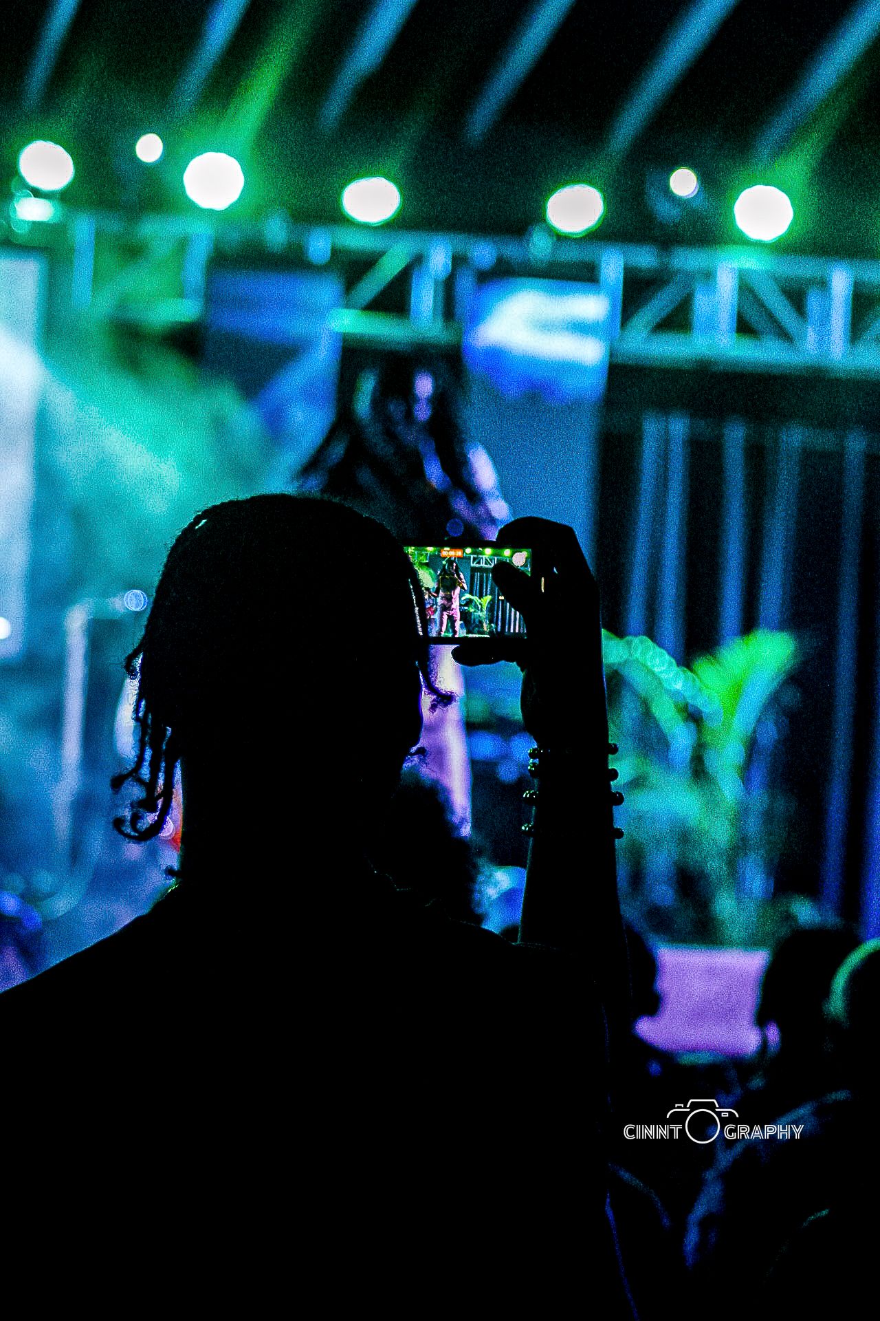Silhouetted concert attendee holds up phone to record performance with blue and green stage lights in background.