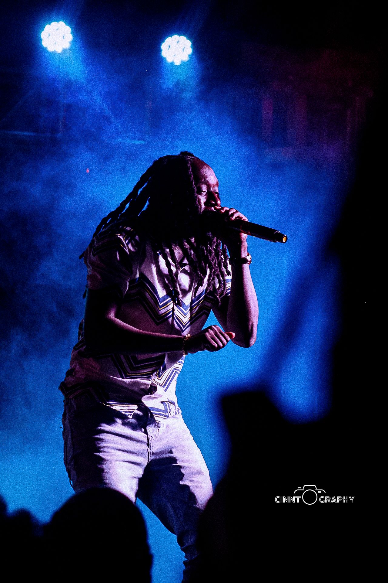 Singer performs on stage with microphone under dramatic blue concert lighting and fog effects.