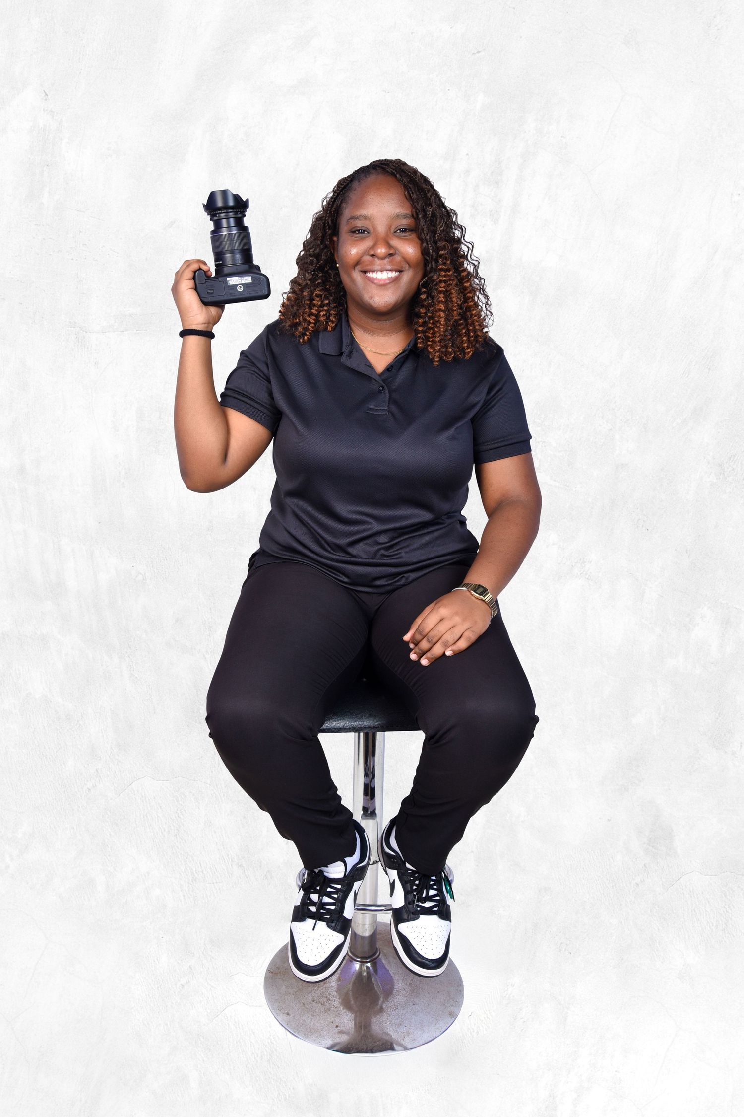 Healthcare worker in dark blue scrubs and athletic shoes smiles while seated on stool against white background.