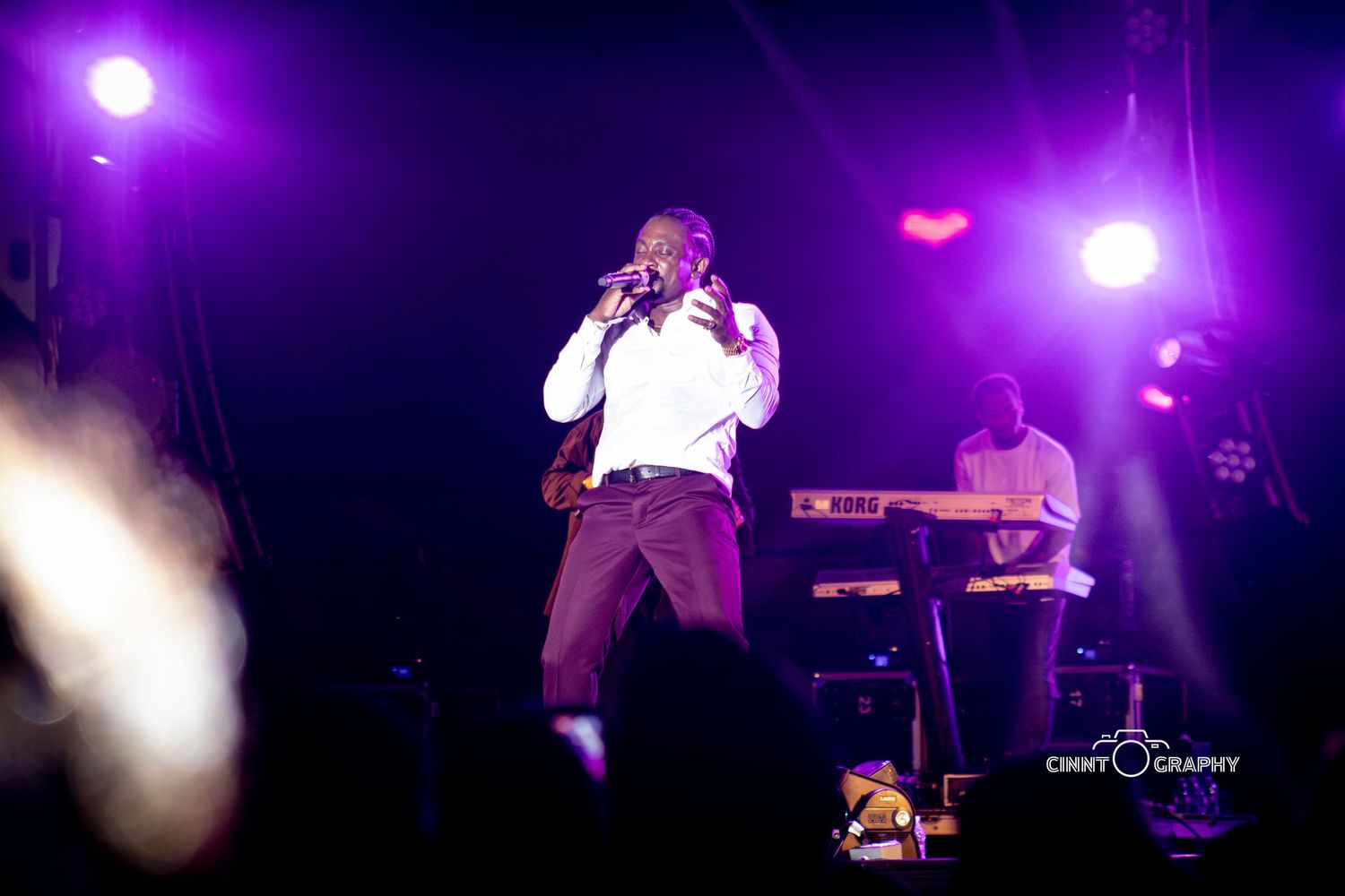 A performer energetically sings on stage illuminated by vibrant purple lighting during a live concert performance.