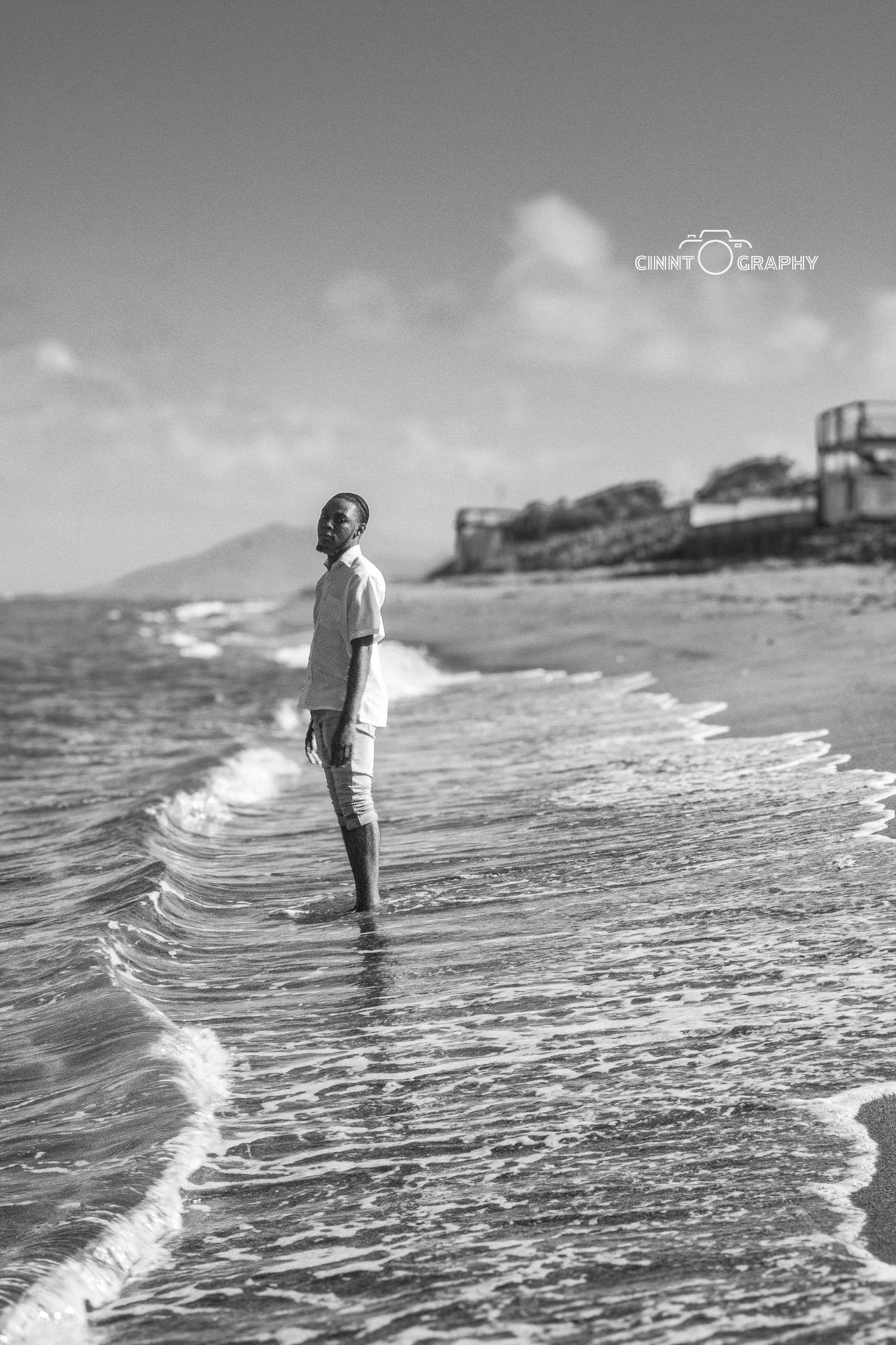 A person stands thoughtfully in shallow ocean waves at the shoreline in a striking black and white photograph.