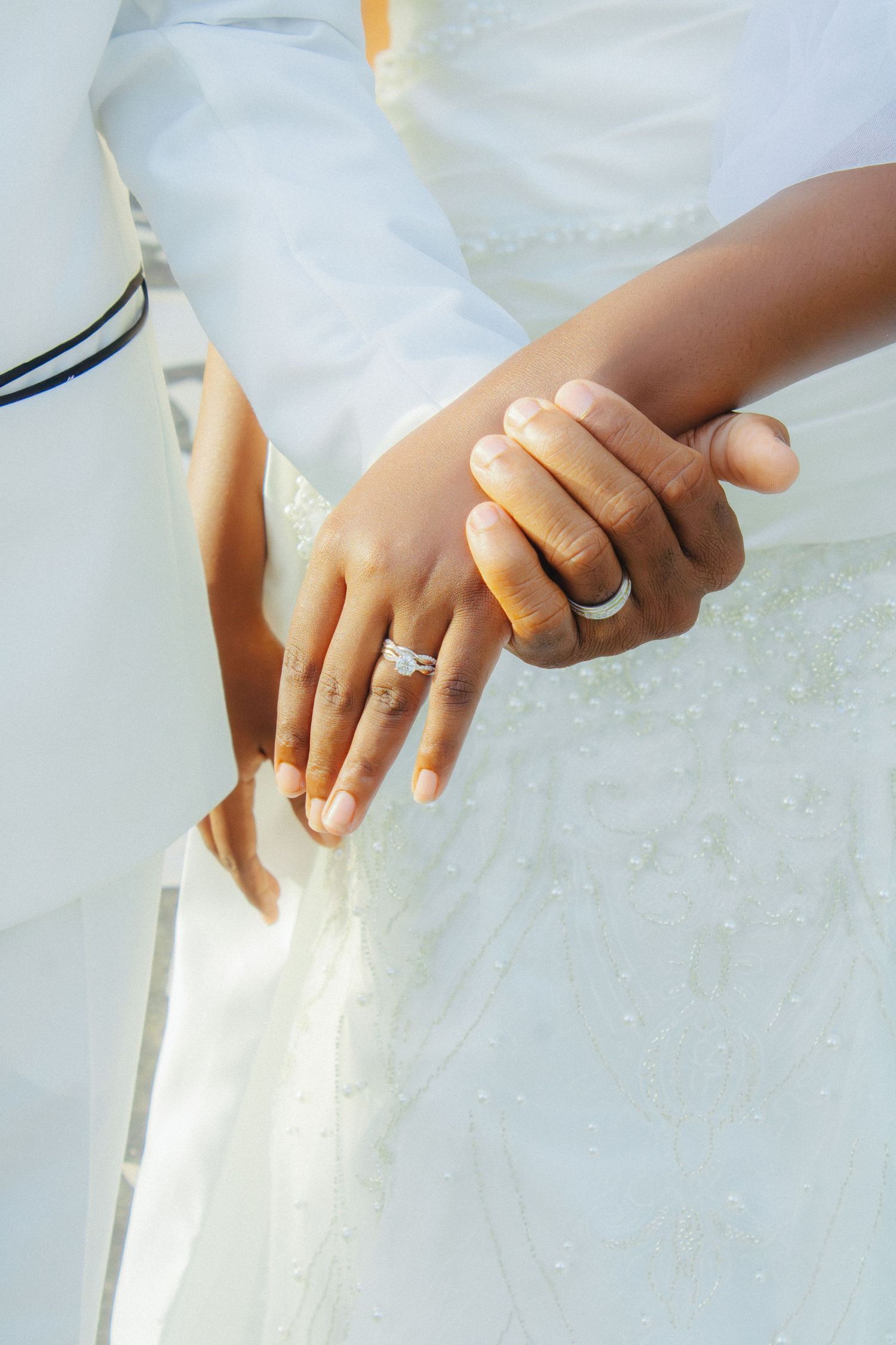 Bride and groom holding hands showing wedding rings, wearing white wedding attire, close-up detail shot.