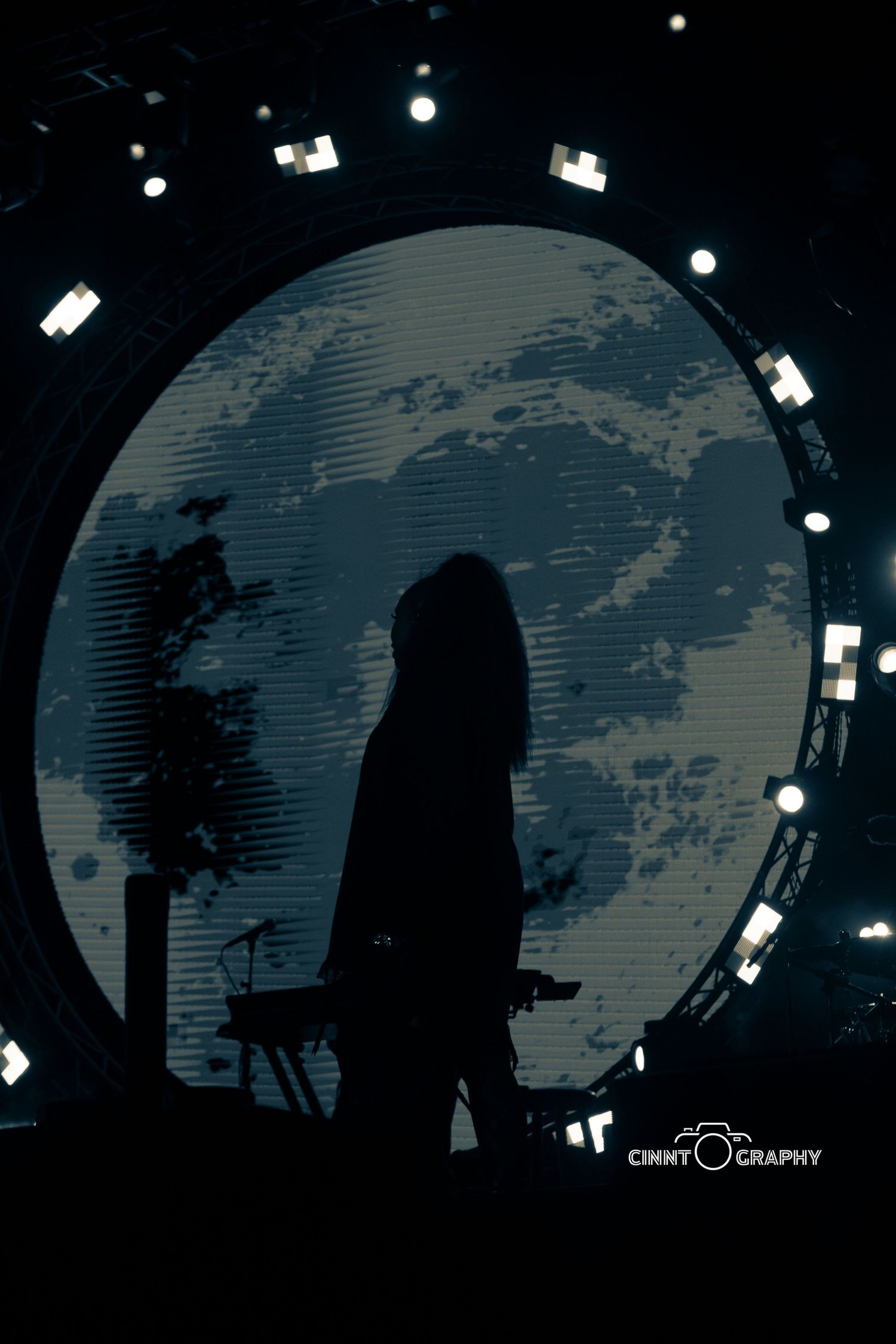 A silhouetted figure stands against a circular backdrop with stage lighting creating a dramatic moonlit effect.