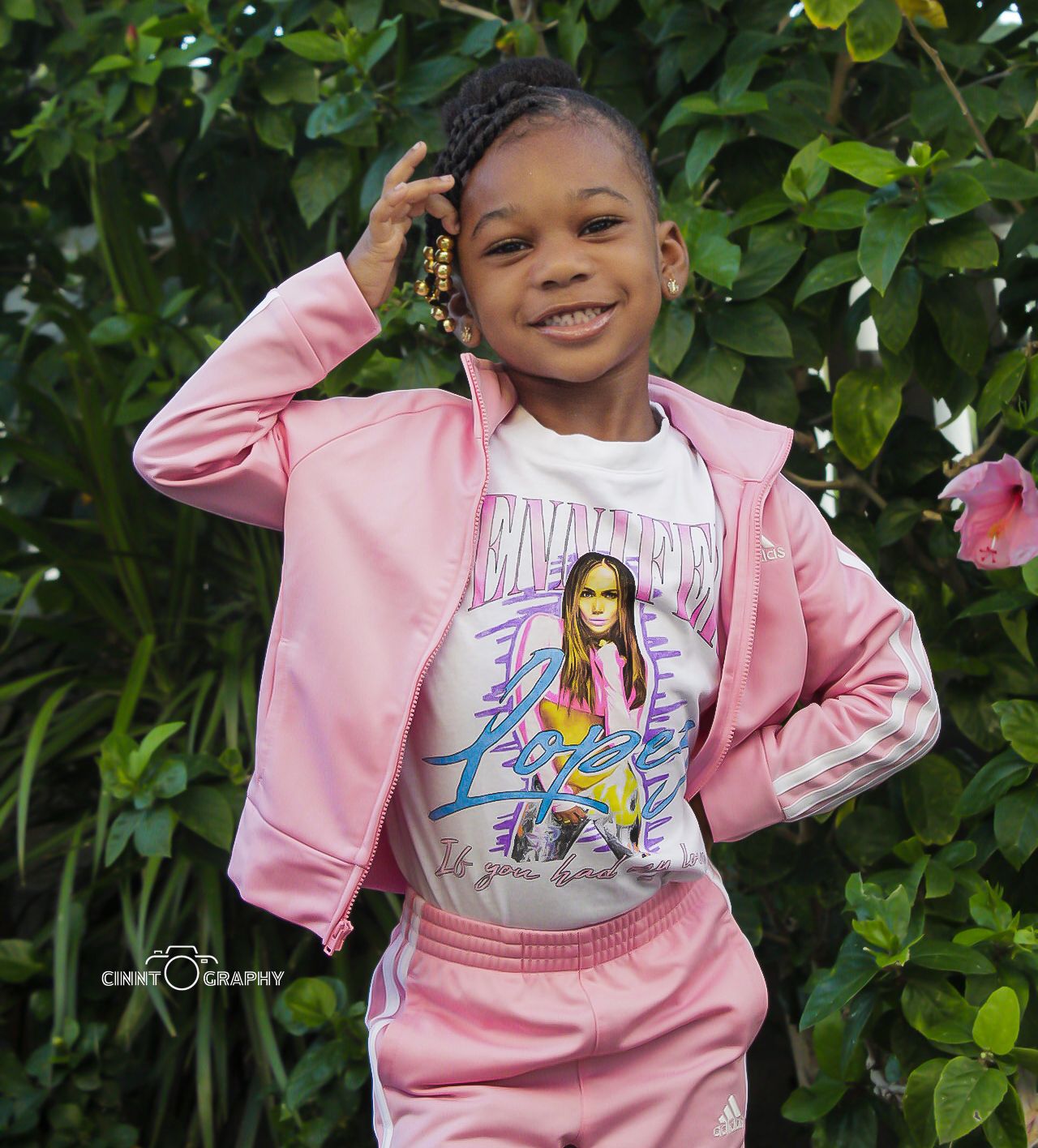 Child wearing a pink tracksuit and graphic t-shirt poses confidently against green foliage in an outdoor setting.