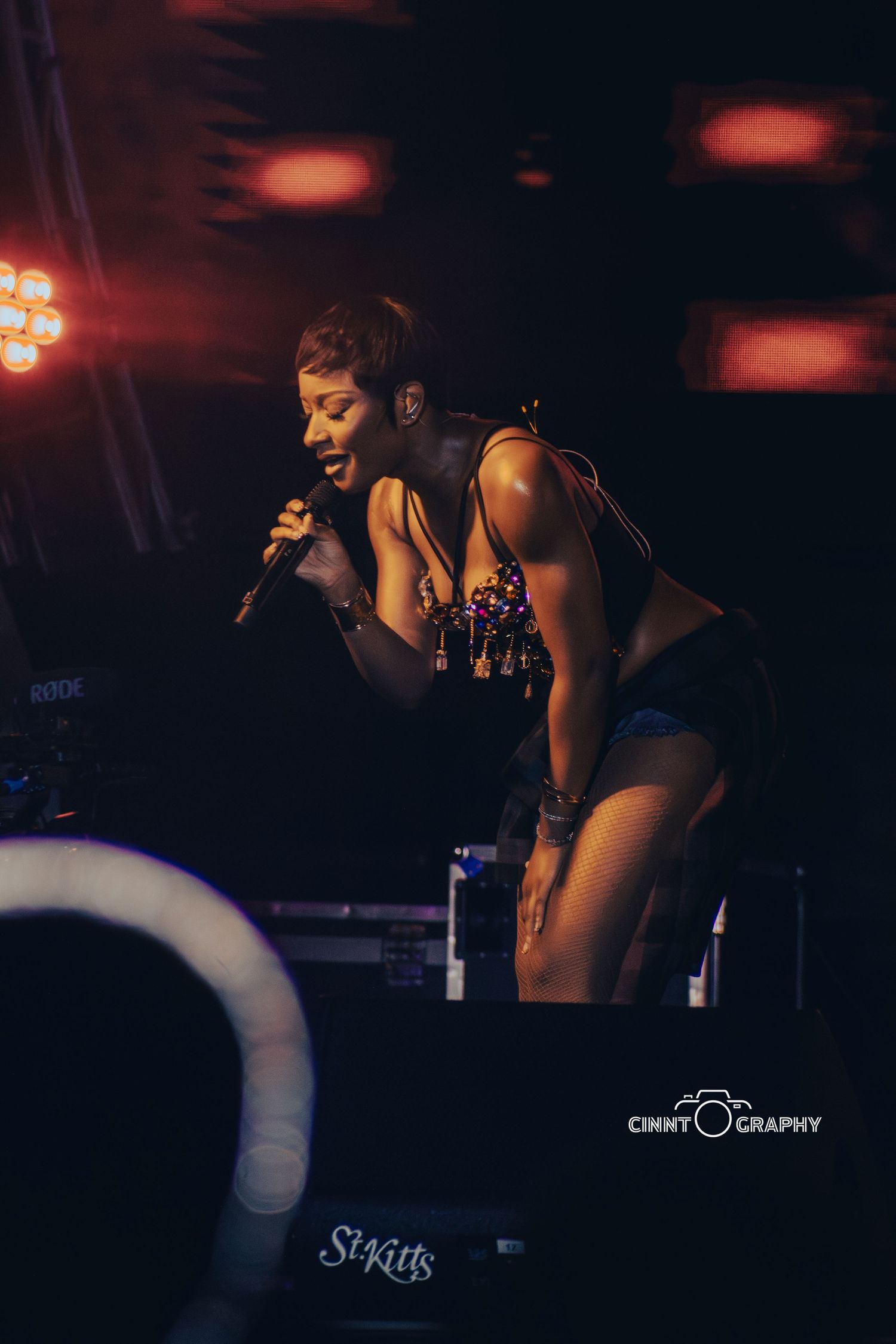 A performer in a black outfit sings into a microphone on a dimly lit stage with red lighting.