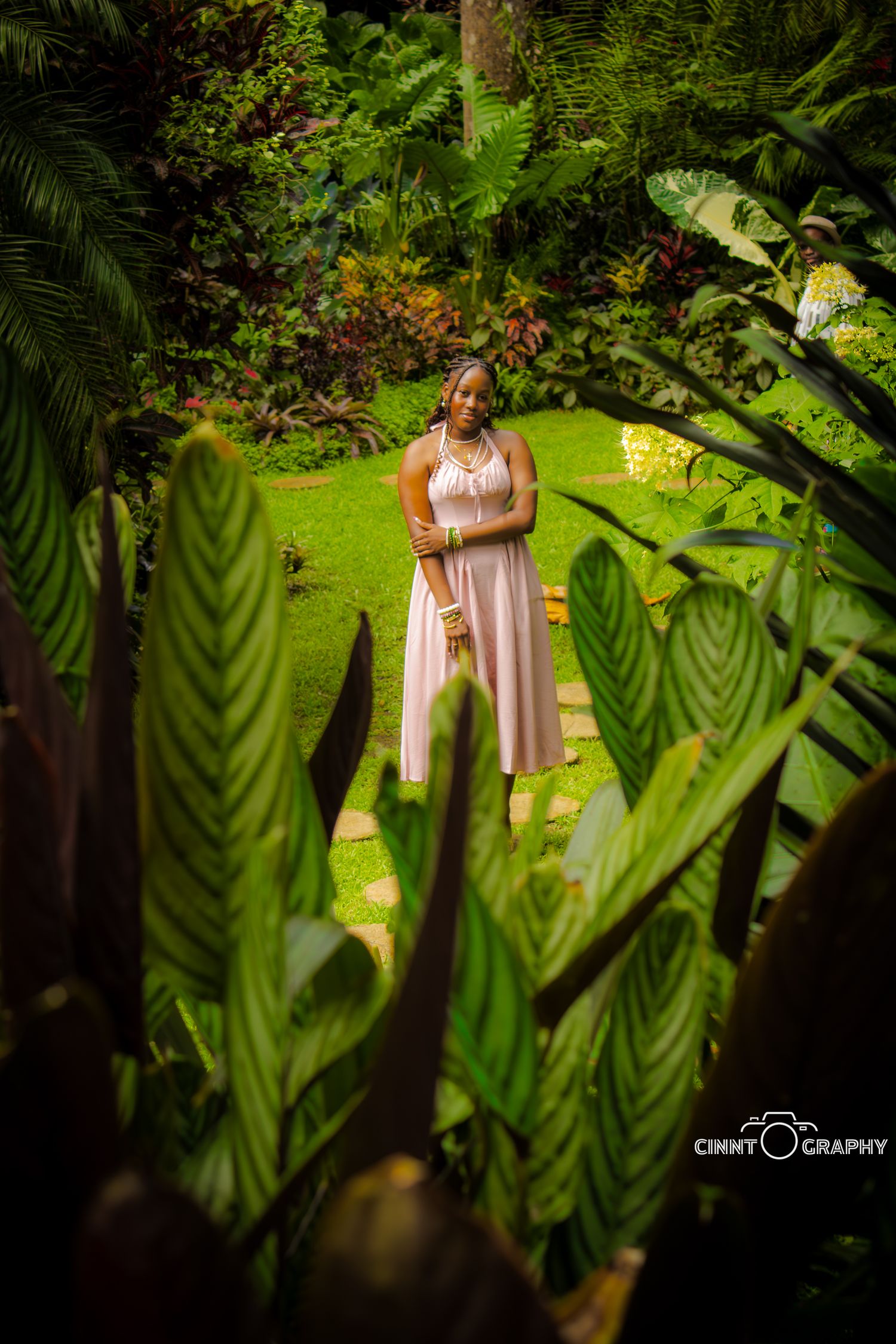 A figure in a flowing beige dress stands in a lush tropical garden framed by large green leaves and foliage.