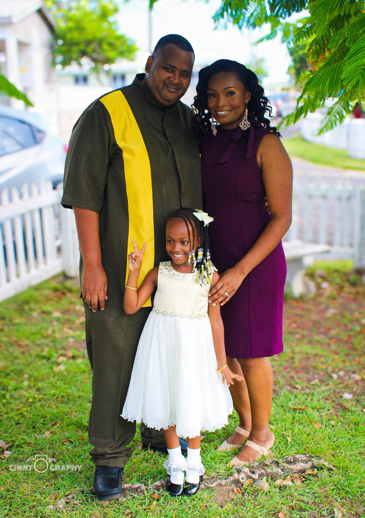 Family of three stands together outdoors in formal attire for a special occasion against a white picket fence and greenery.