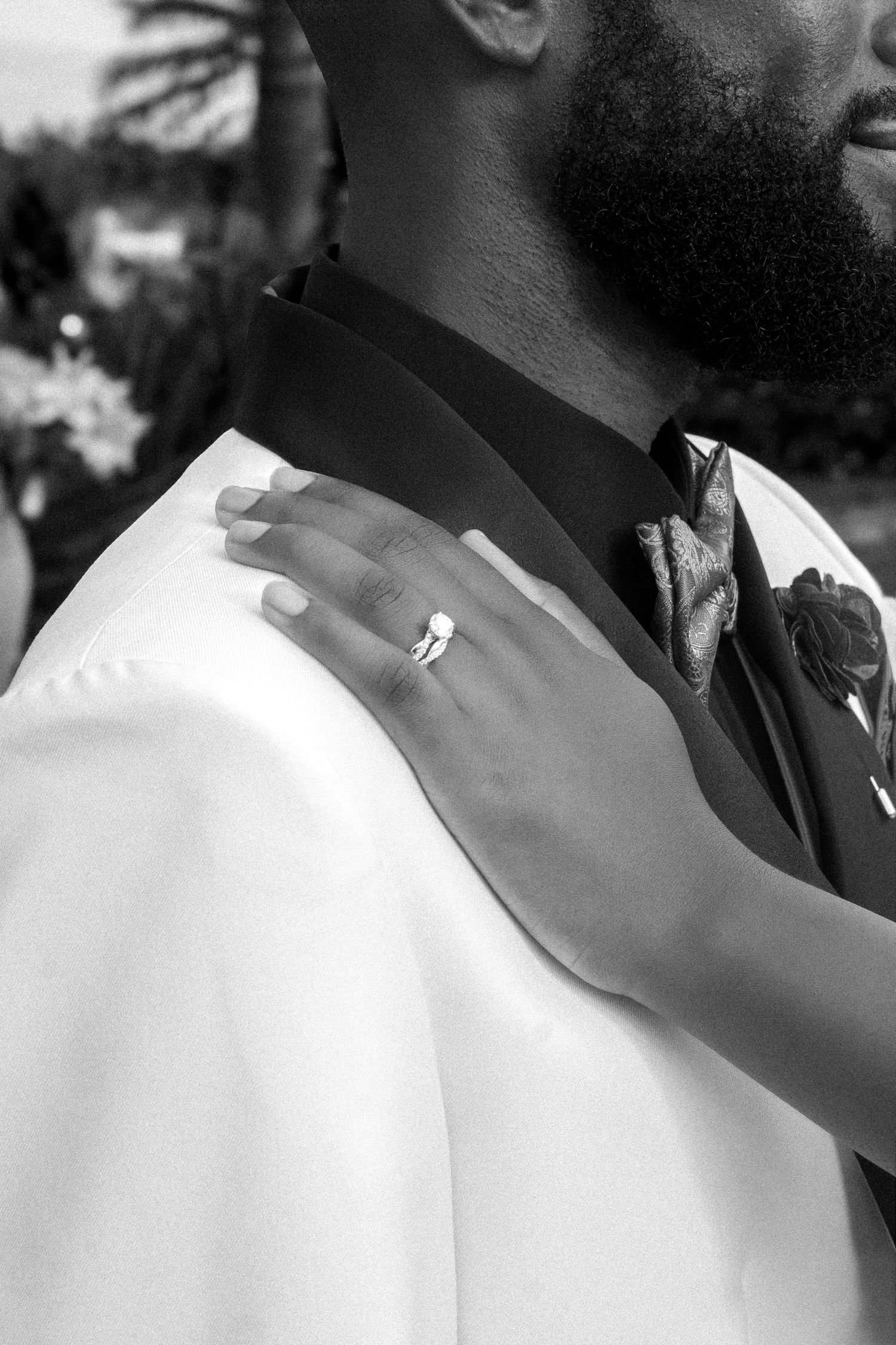 Black and white photo of a bride's hand with engagement ring resting on groom's white tuxedo shoulder.