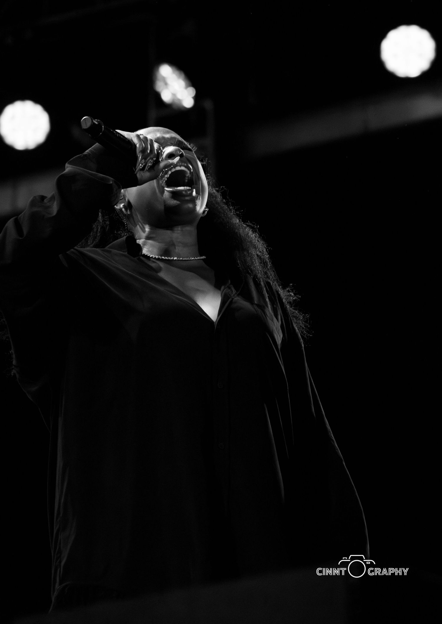 Black and white concert photo of a performer singing passionately with stage lights in dark background.