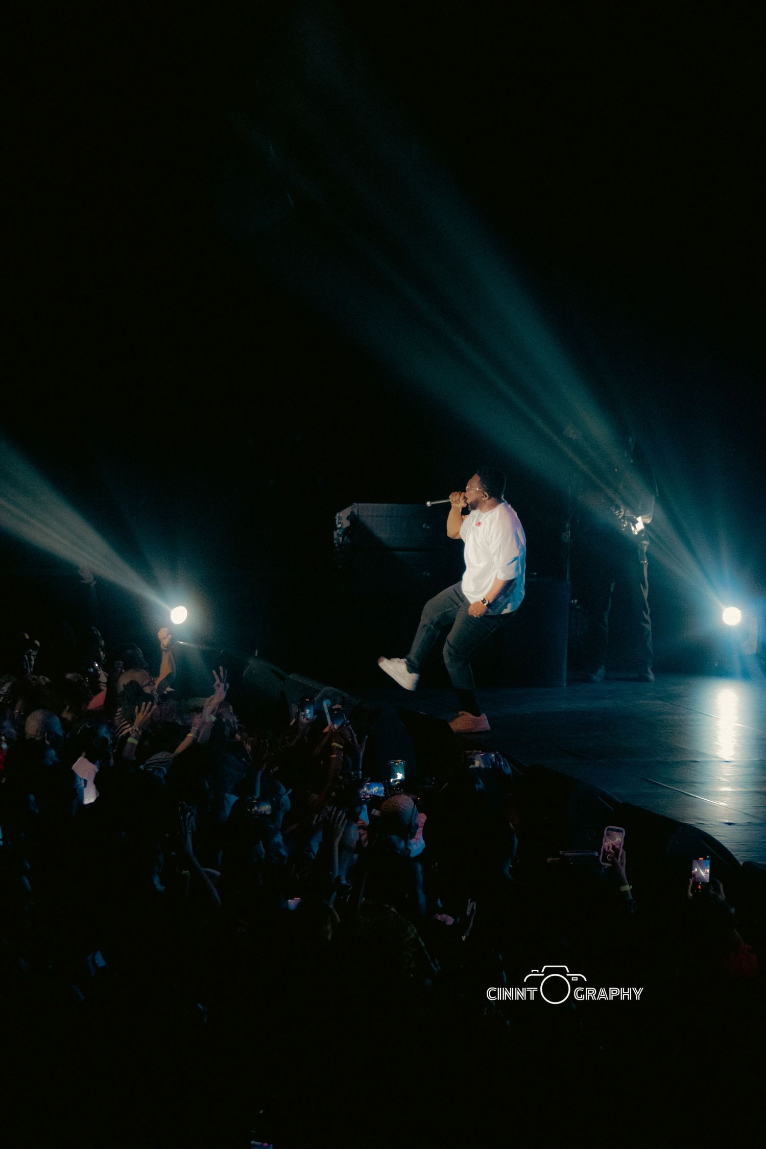 Performer leaps high in the air during a concert with dramatic blue stage lighting and silhouetted crowd below.