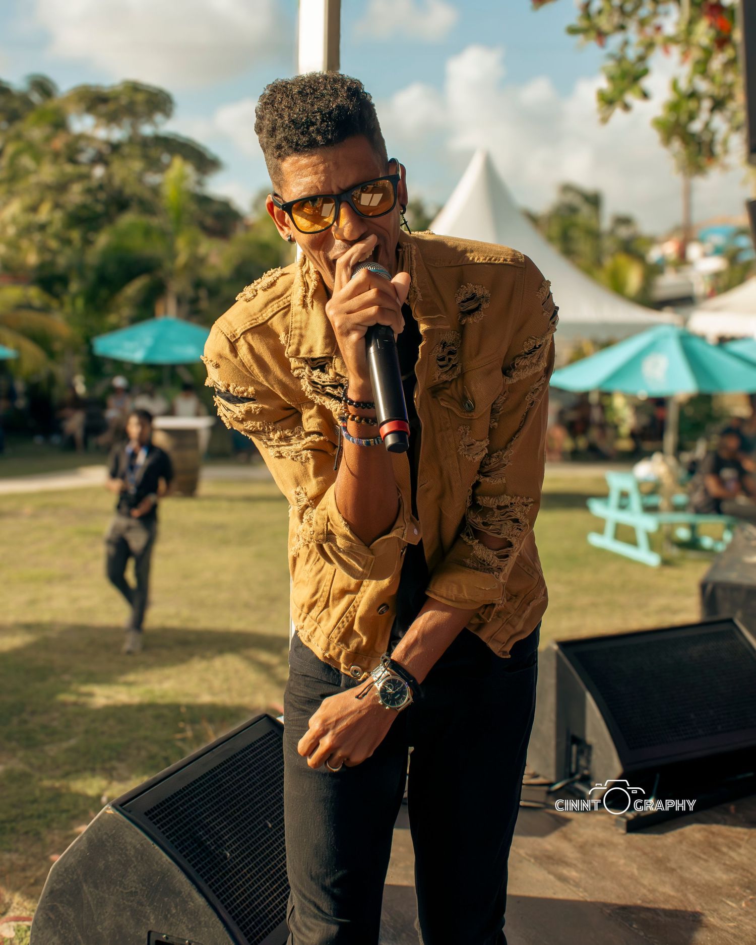 Performer in a brown jacket and glasses stands on an outdoor stage with umbrellas and trees visible in the background.