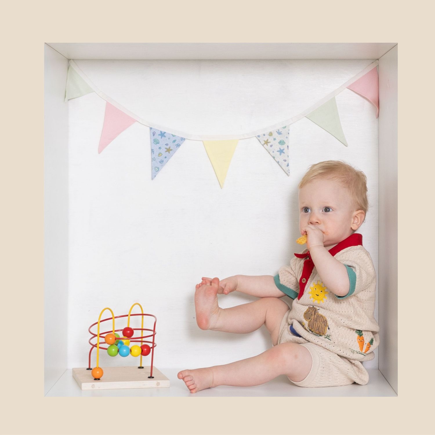 Cute baby sitting in a white box frame with colorful bunting flags and a wooden abacus toy.