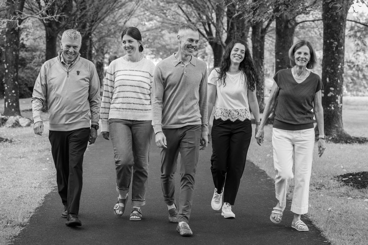 Black and white photo of five adults walking together on a tree-lined path outdoors.