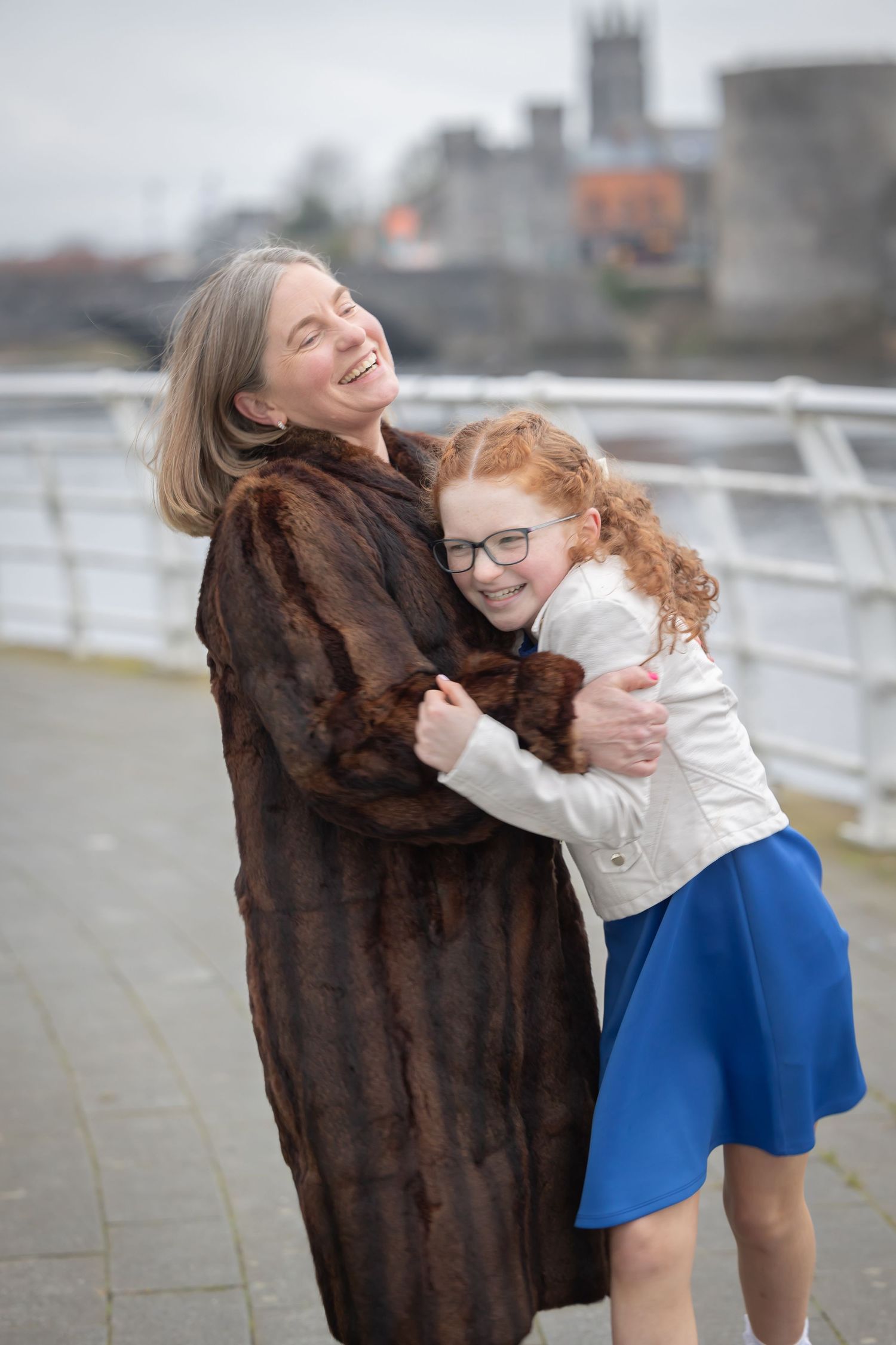 A joyful moment shared between two people embracing and laughing together on a cloudy day by the waterfront.