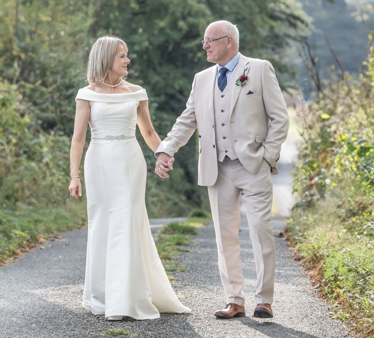 Couple walking hand in hand on path, wearing matching white and beige formal attire.