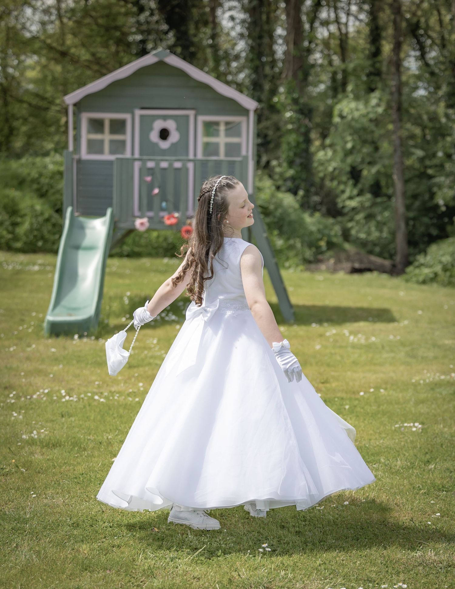 Child in a flowing white dress twirls in a sunny garden with a playhouse in the background.