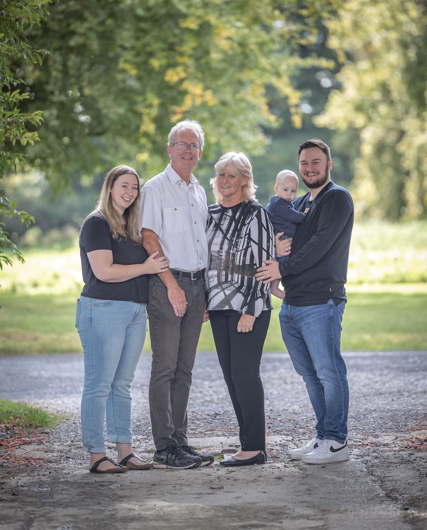 A family group poses together on a gravel path surrounded by lush green trees in a casual outdoor setting.