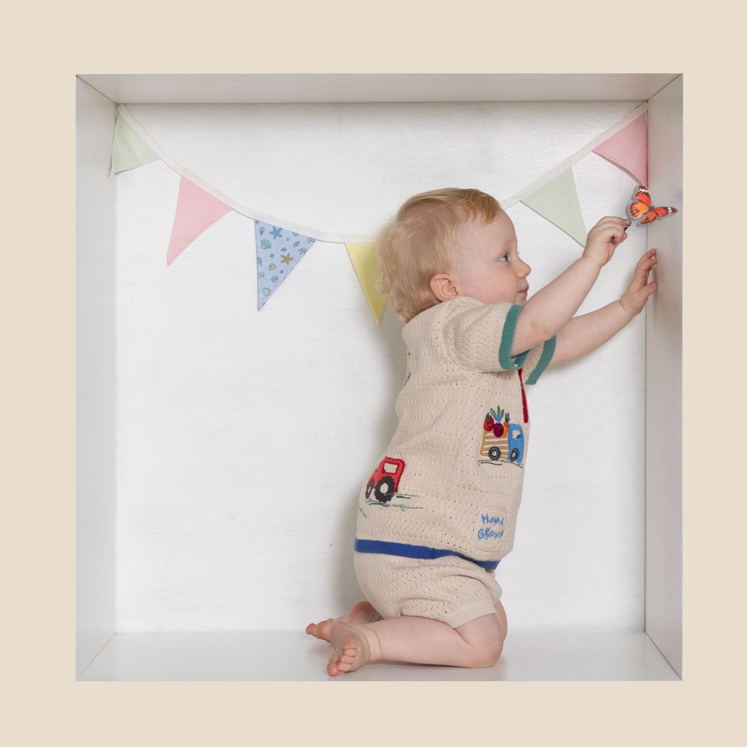 A baby in a romper reaches up to touch colorful bunting flags in a bright white box frame display.