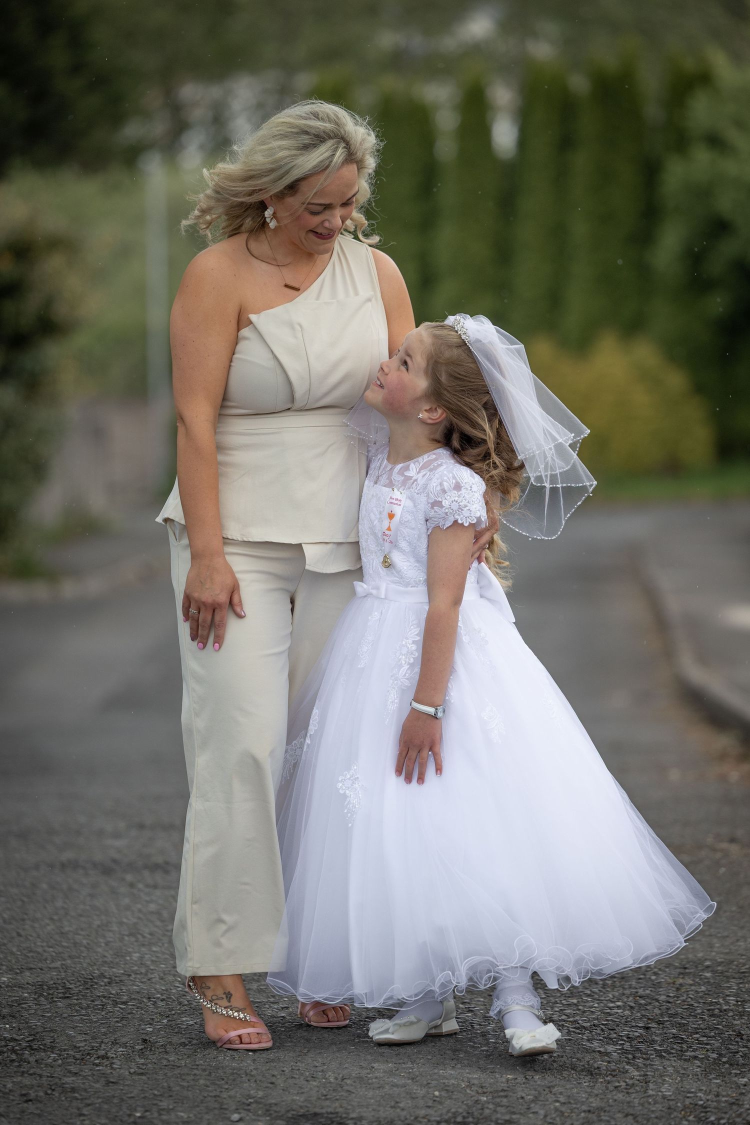 A woman in beige pantsuit smiles at a young girl in white First Communion dress and veil on a paved path.
