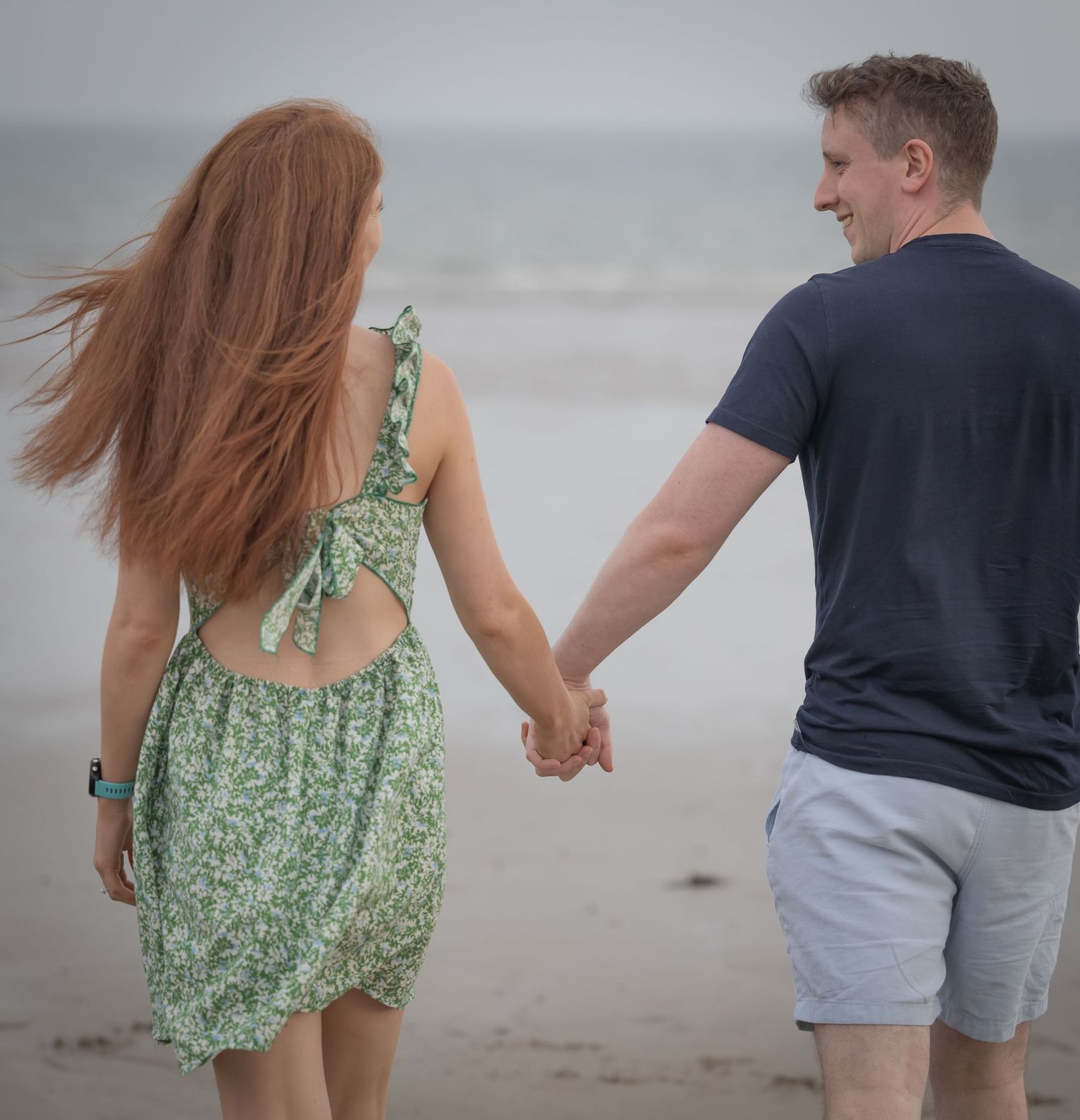 Couple holding hands walking on a beach, woman in green floral dress with red hair, man in navy t-shirt and gray shorts.