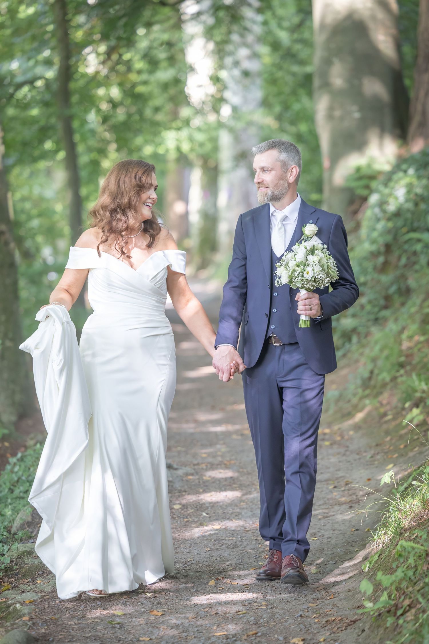 Couple walking hand in hand on forest path, bride in white dress and groom in navy suit.